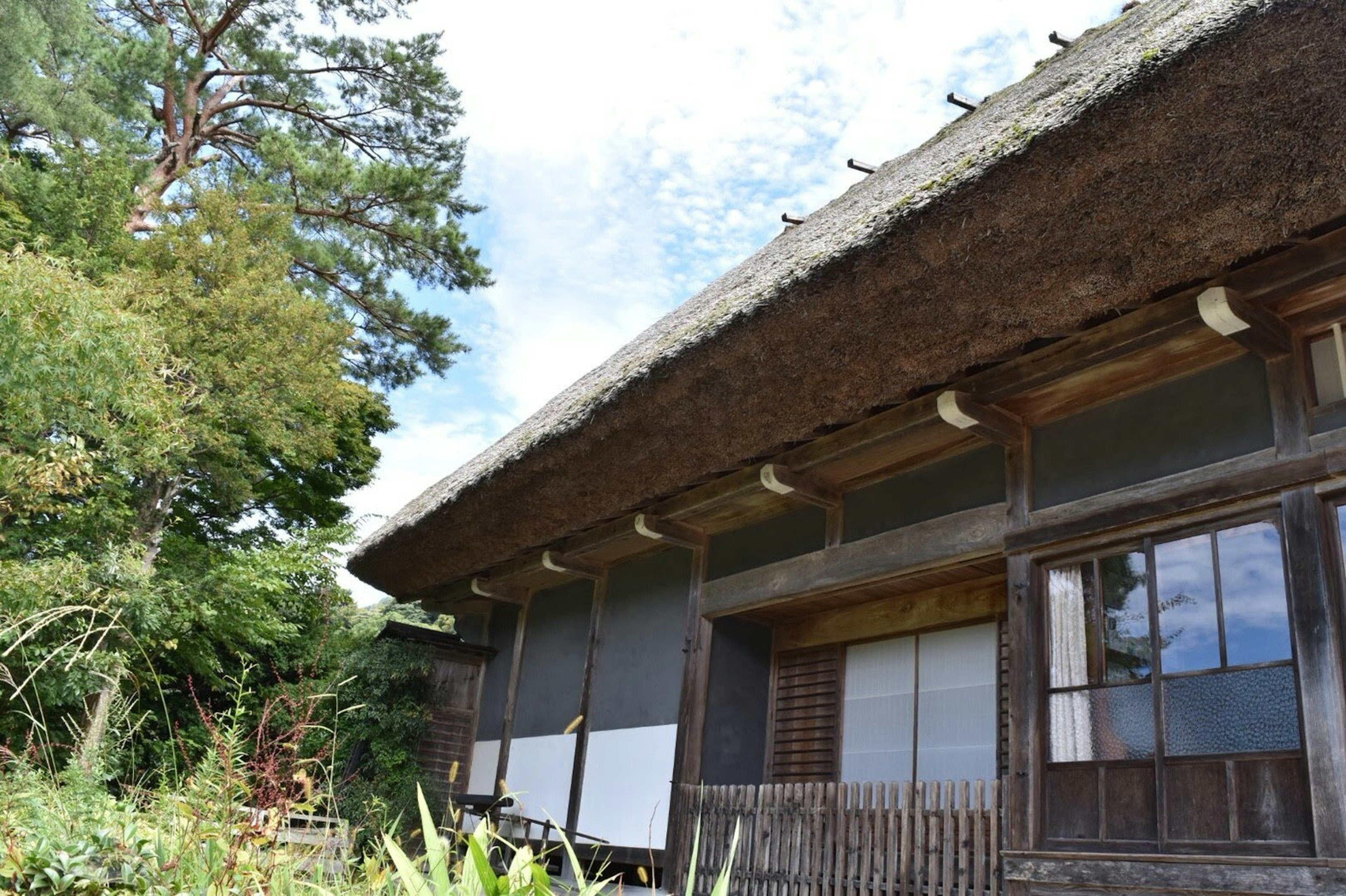 Eksterior rumah Jepang tradisional dikelilingi pohon hijau dengan atap jerami dan jendela kayu