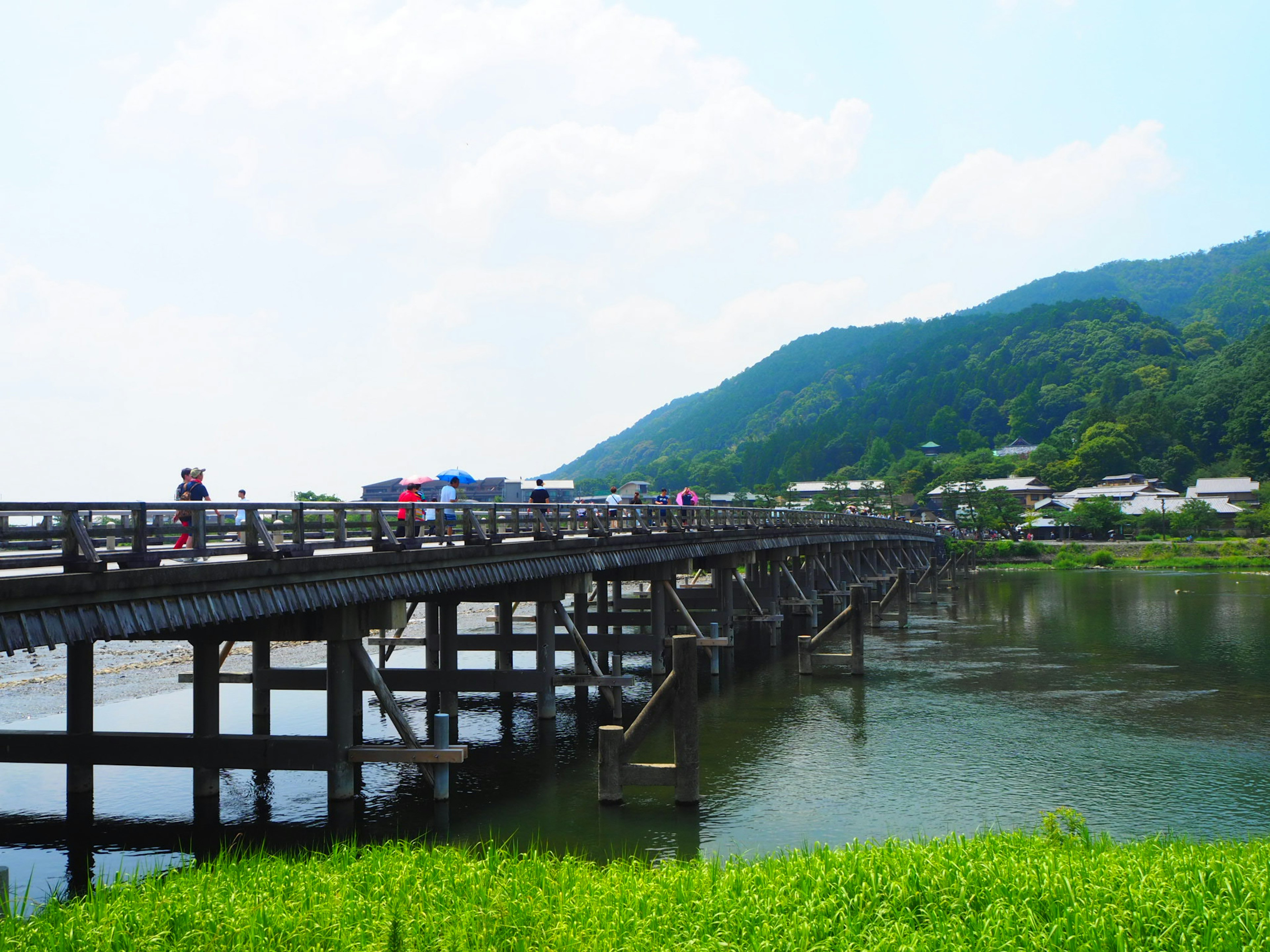 สะพานไม้ข้ามแม่น้ำที่มีผู้คนเดินและภูเขาสีเขียว