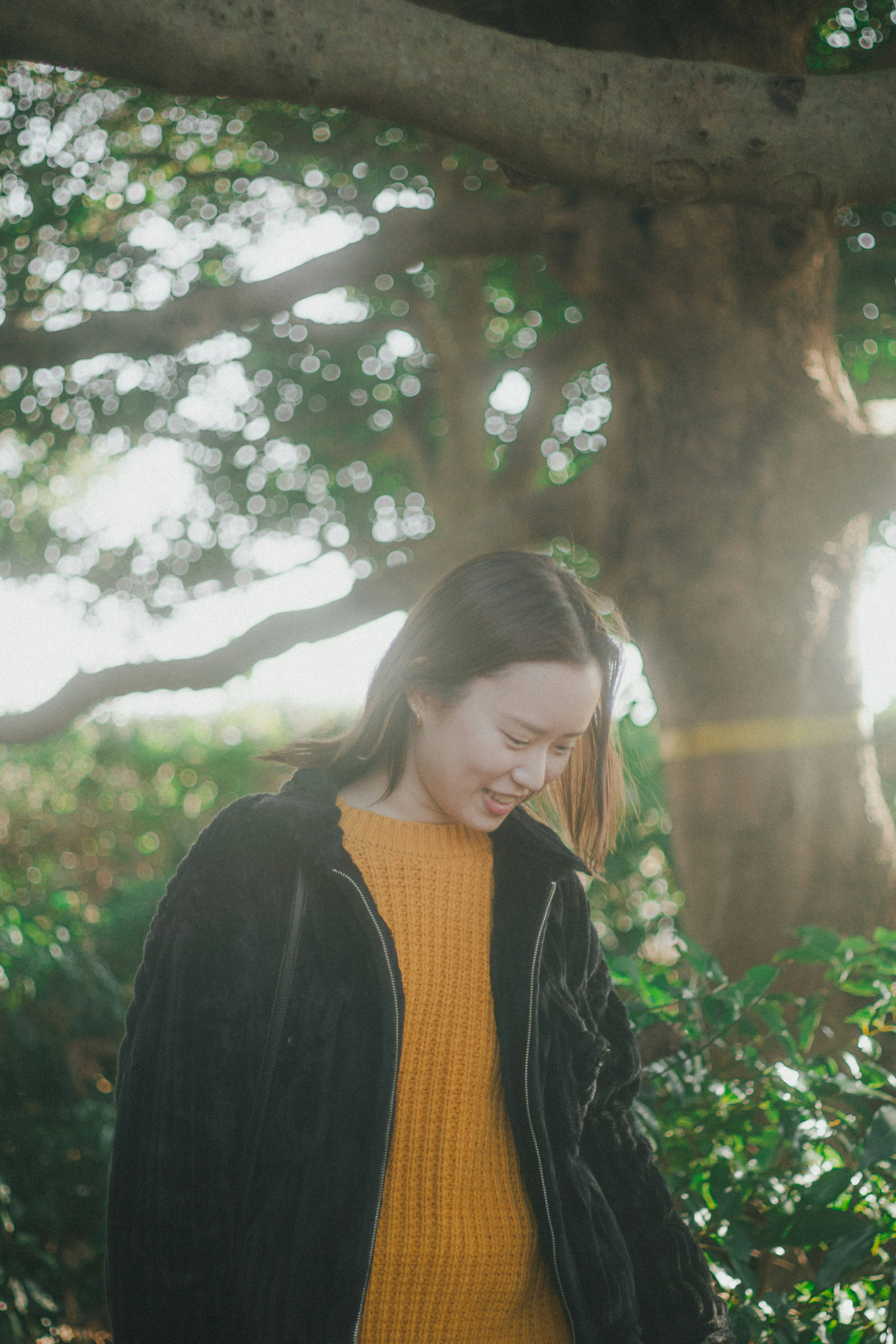 Una donna che indossa un maglione giallo in piedi tra il fogliame verde con la luce del sole che filtra tra gli alberi