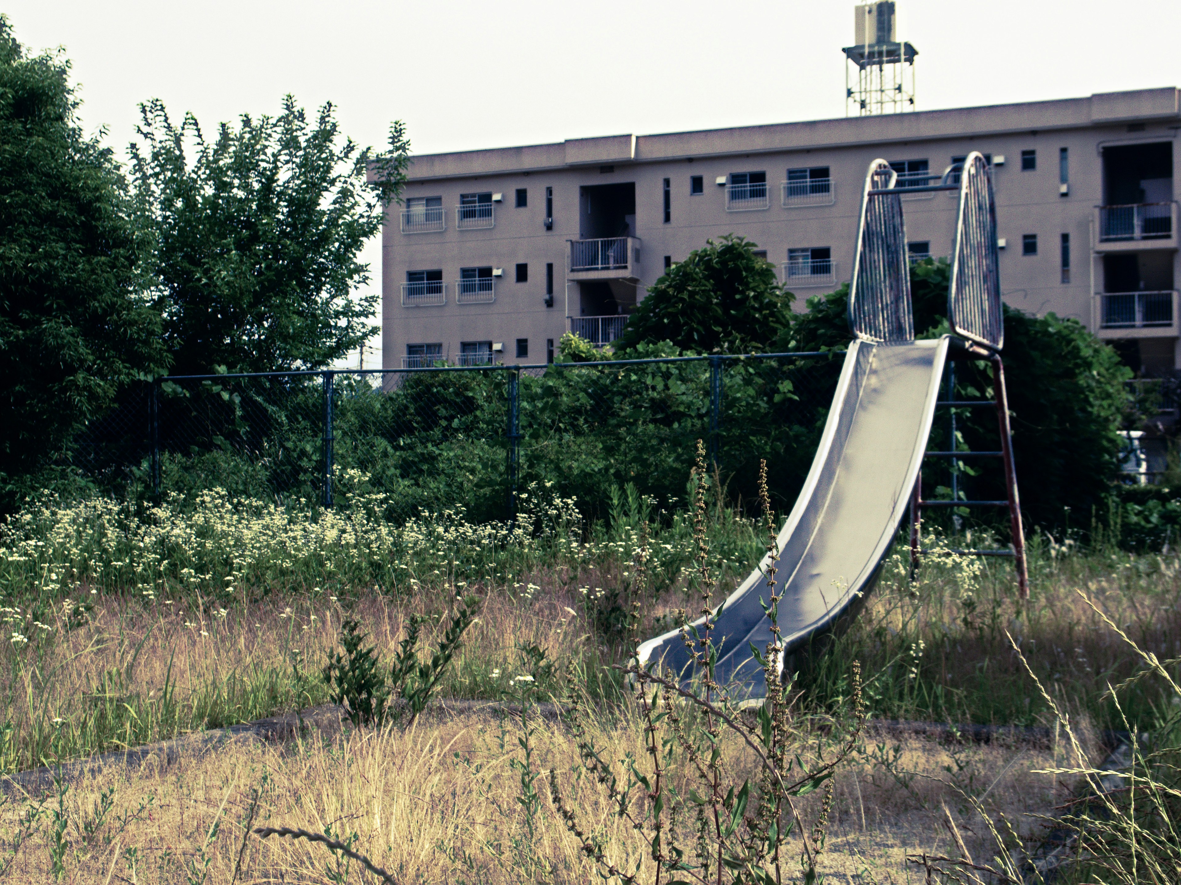 An old slide in a grassy vacant lot with an apartment building in the background