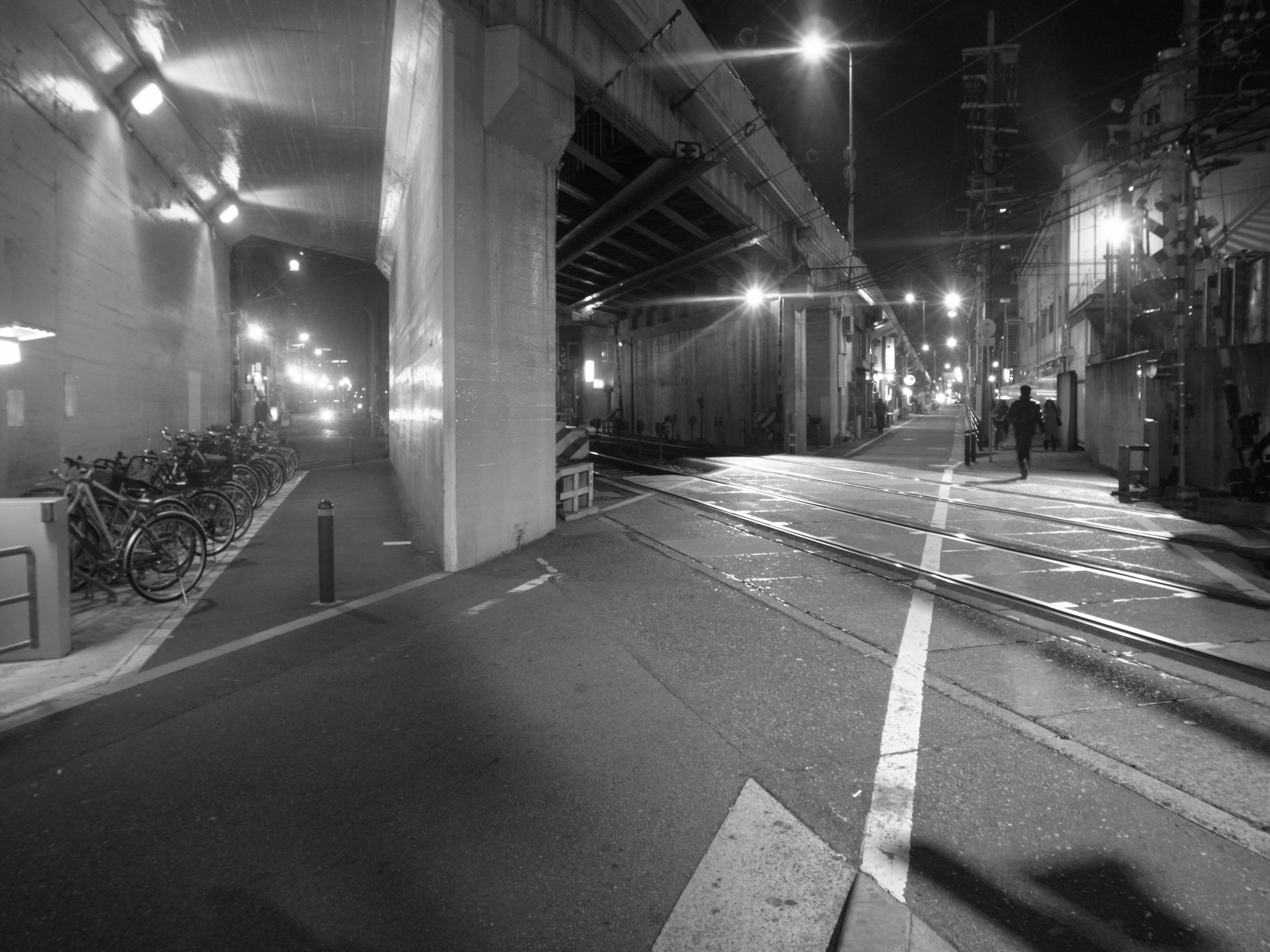 Night cityscape featuring a road under a bridge with bicycle parking and pedestrians