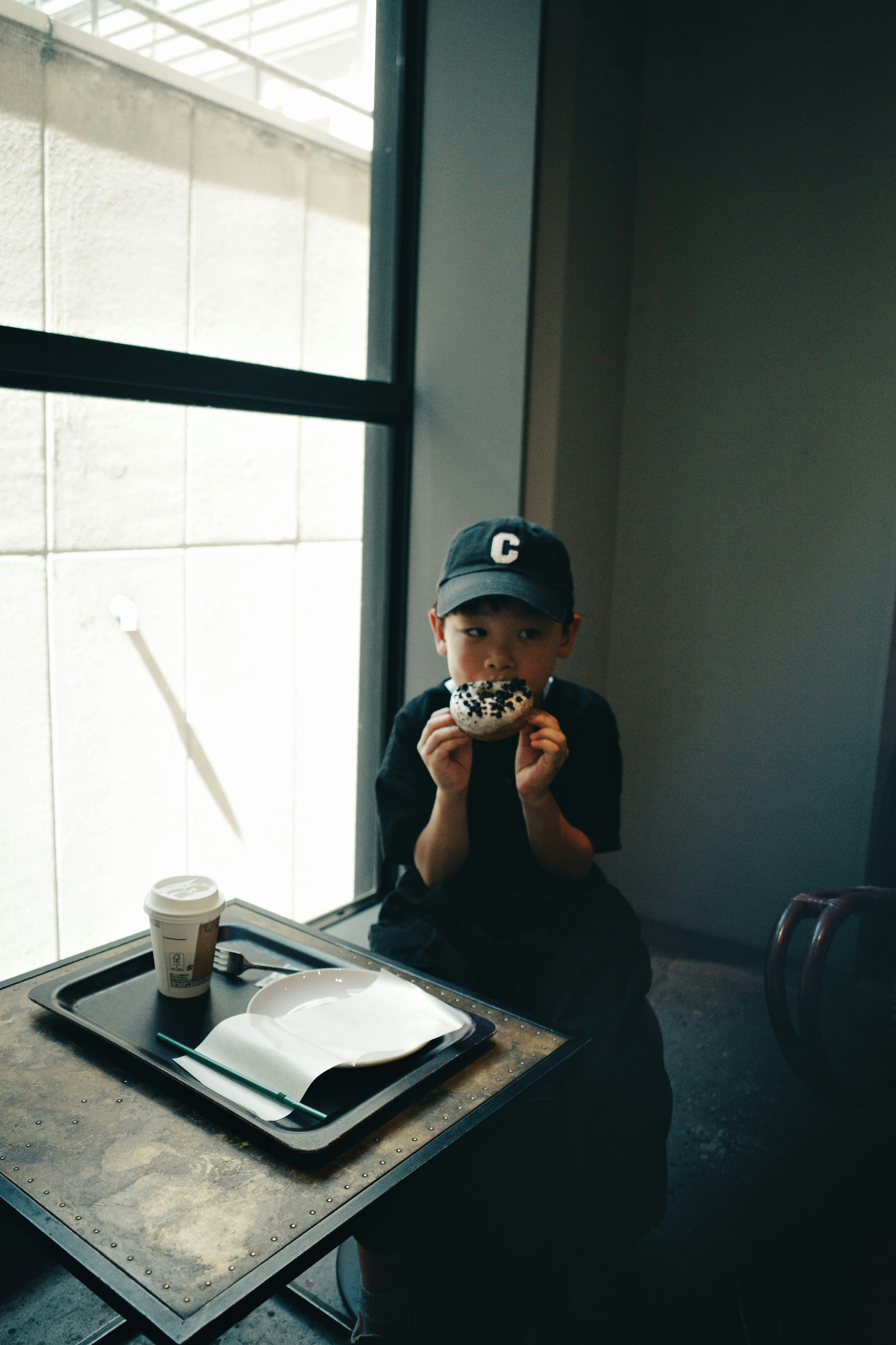 Anak duduk di dekat jendela memegang kamera dengan kopi dan buku catatan di meja