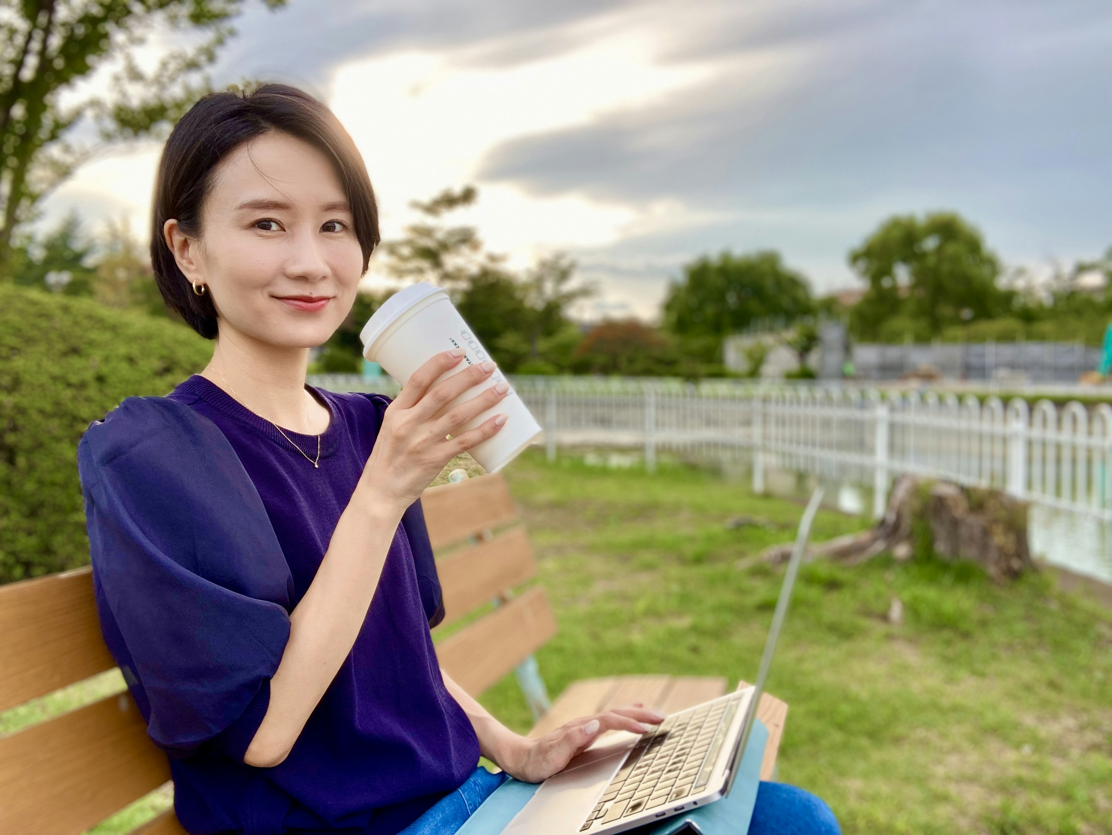 Wanita duduk di bangku taman memegang cangkir kopi sambil bekerja di laptop