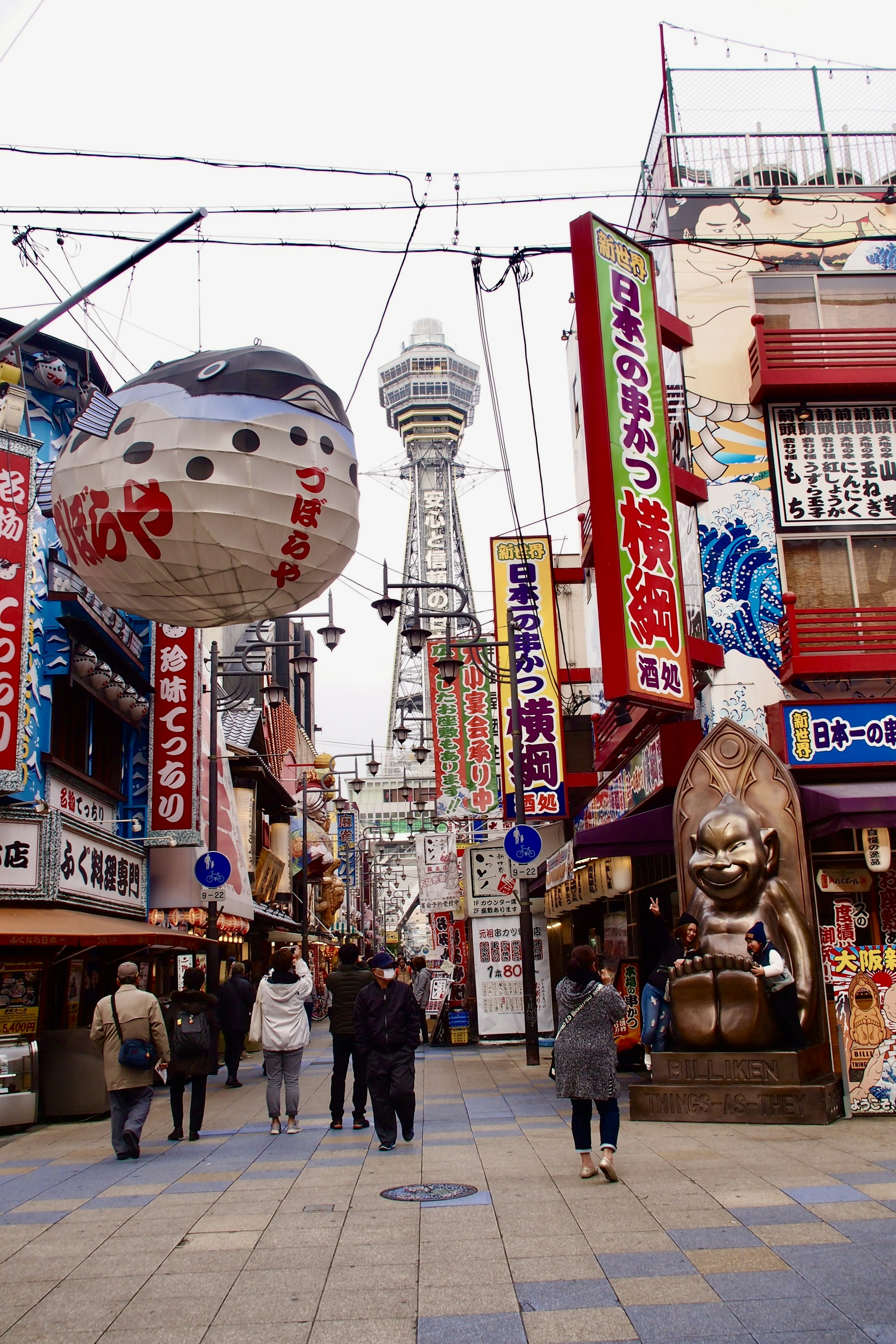 Una calle comercial vibrante con la Torre Tsutenkaku al fondo numerosos letreros y personas caminando