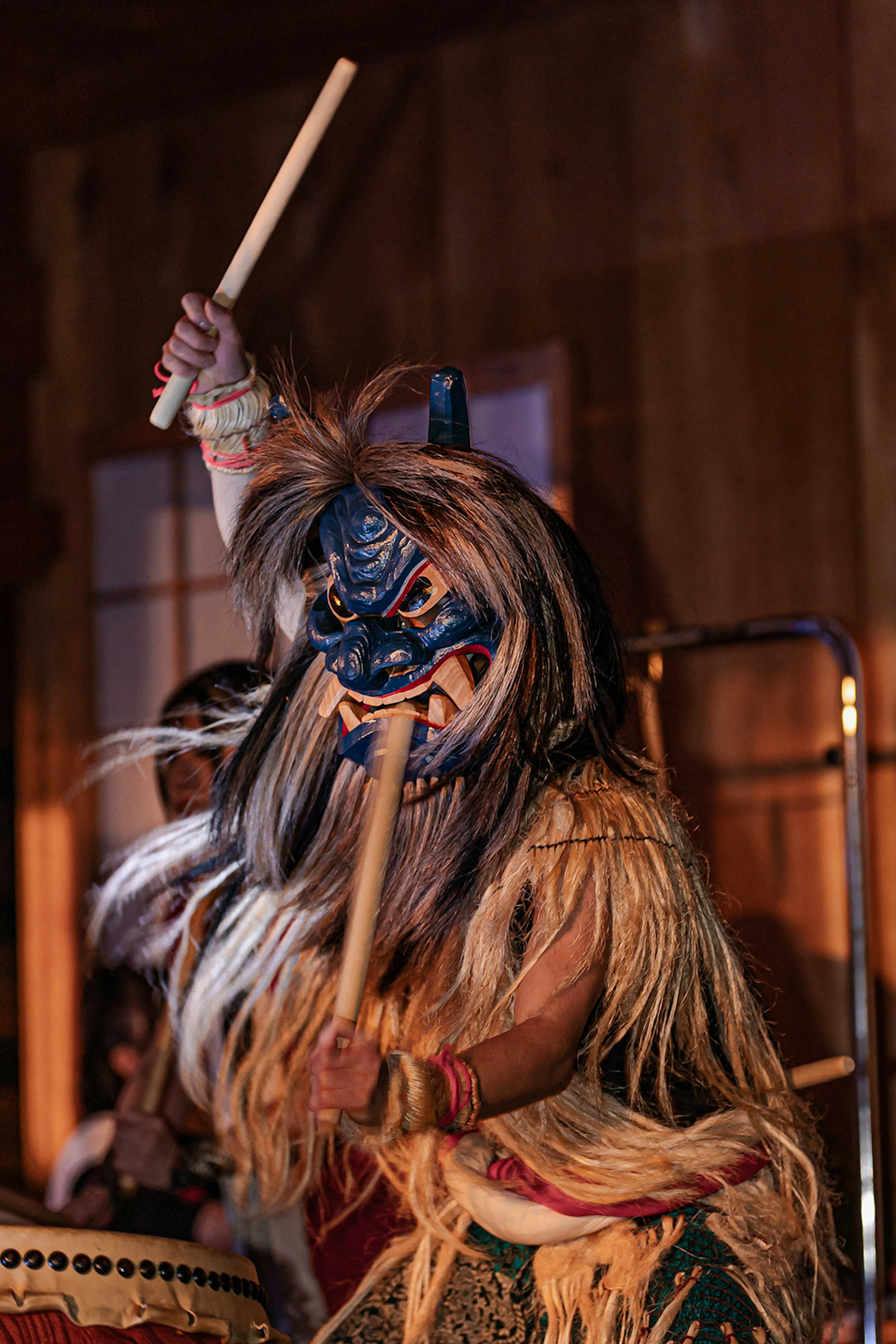 Performer in traditional attire striking a drum with a blue mask and long hair