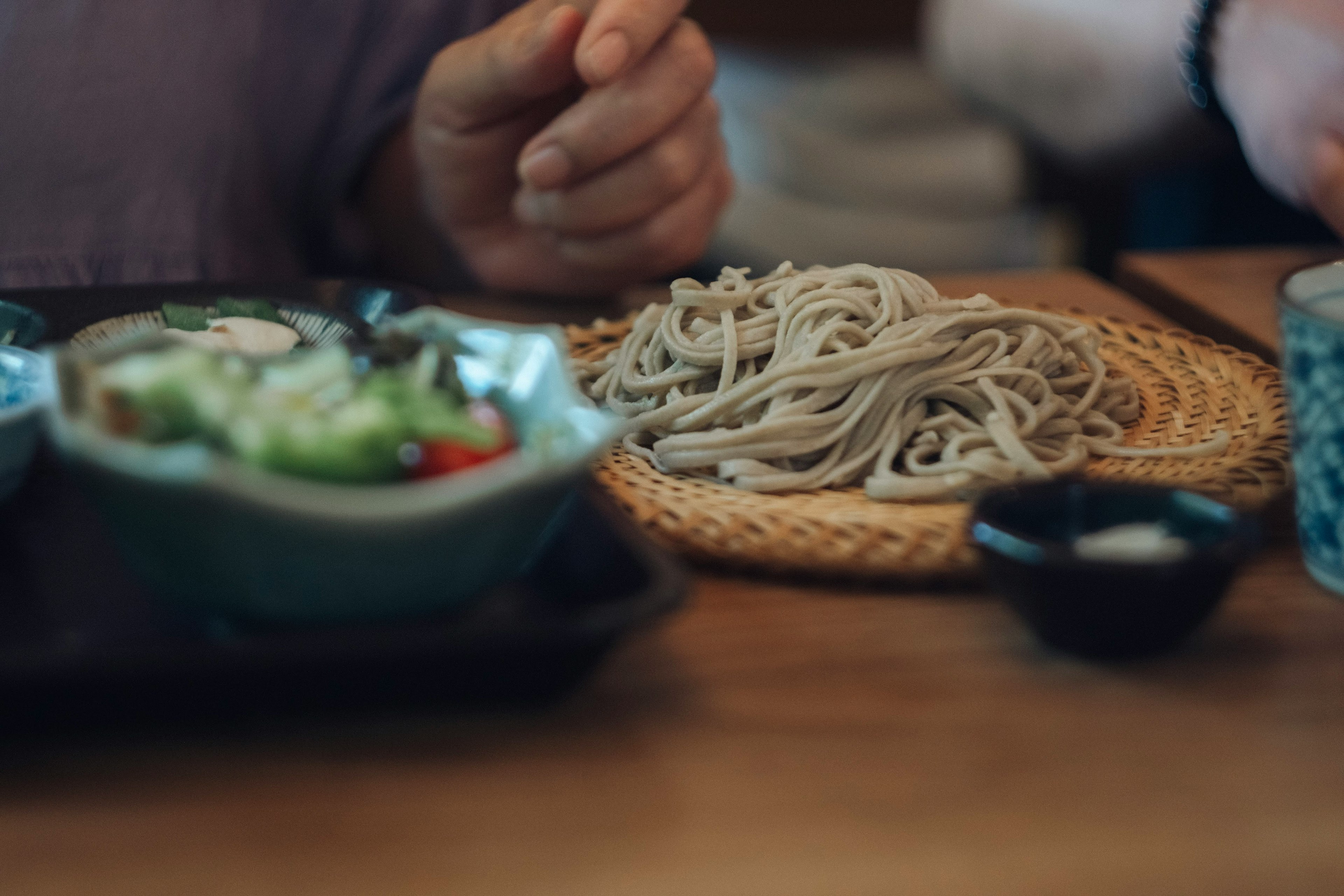 Una persona sosteniendo fideos soba cerca de un plato de soba y ensalada