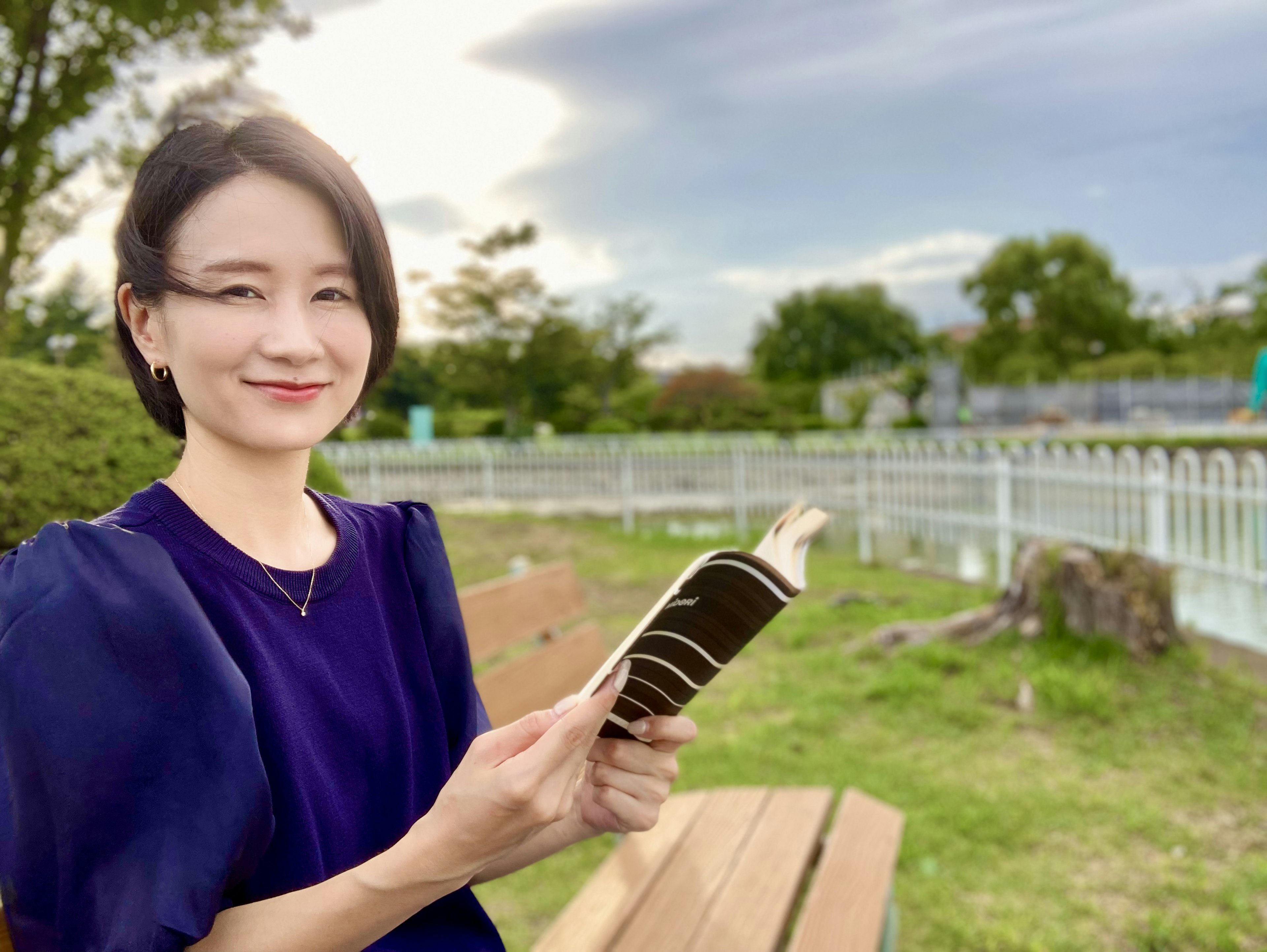 Wanita memegang buku di taman dengan langit cerah dan latar belakang hijau