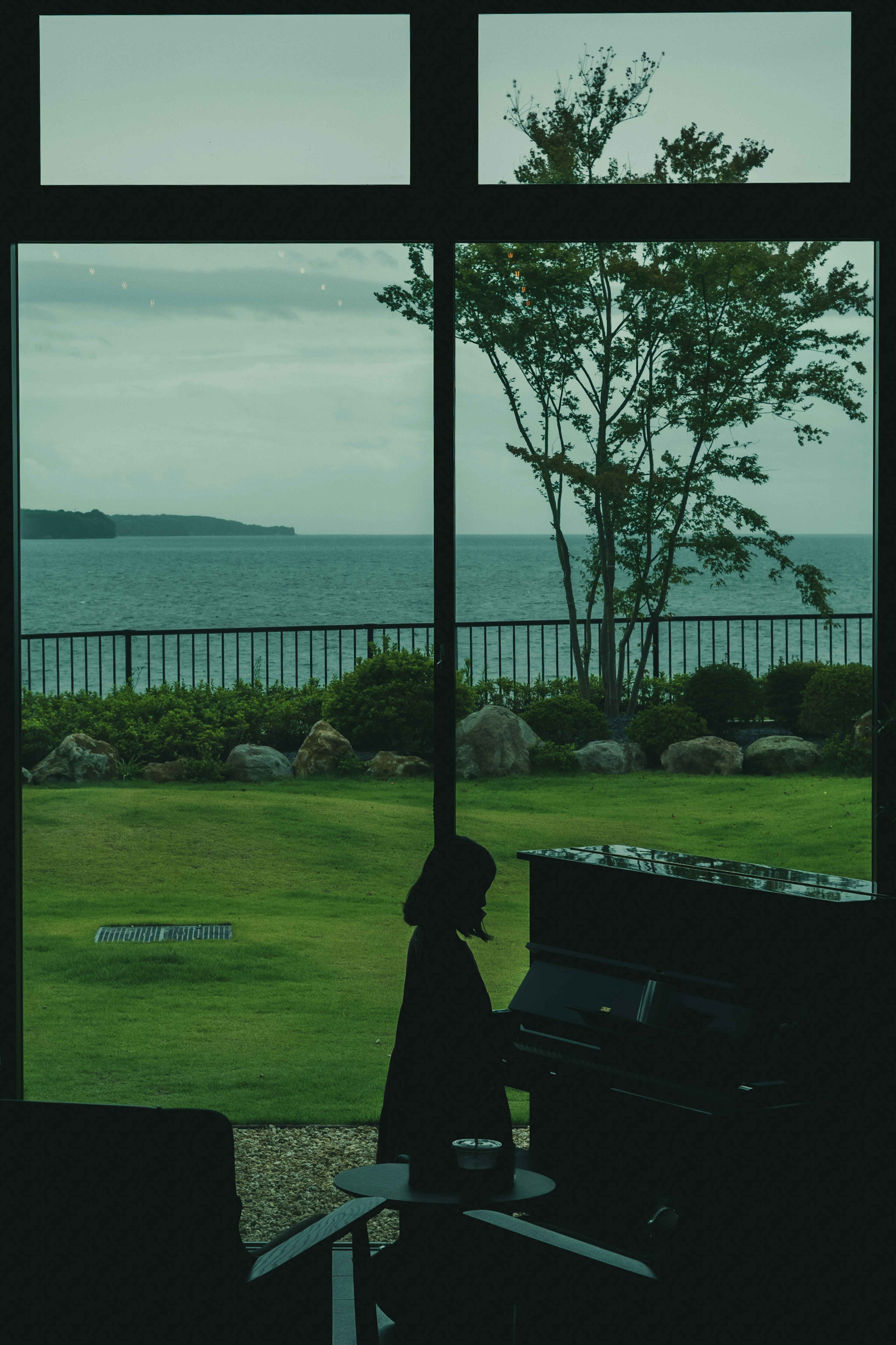 Vista dell'oceano e degli alberi attraverso una finestra con una sedia nera e un pianoforte in primo piano