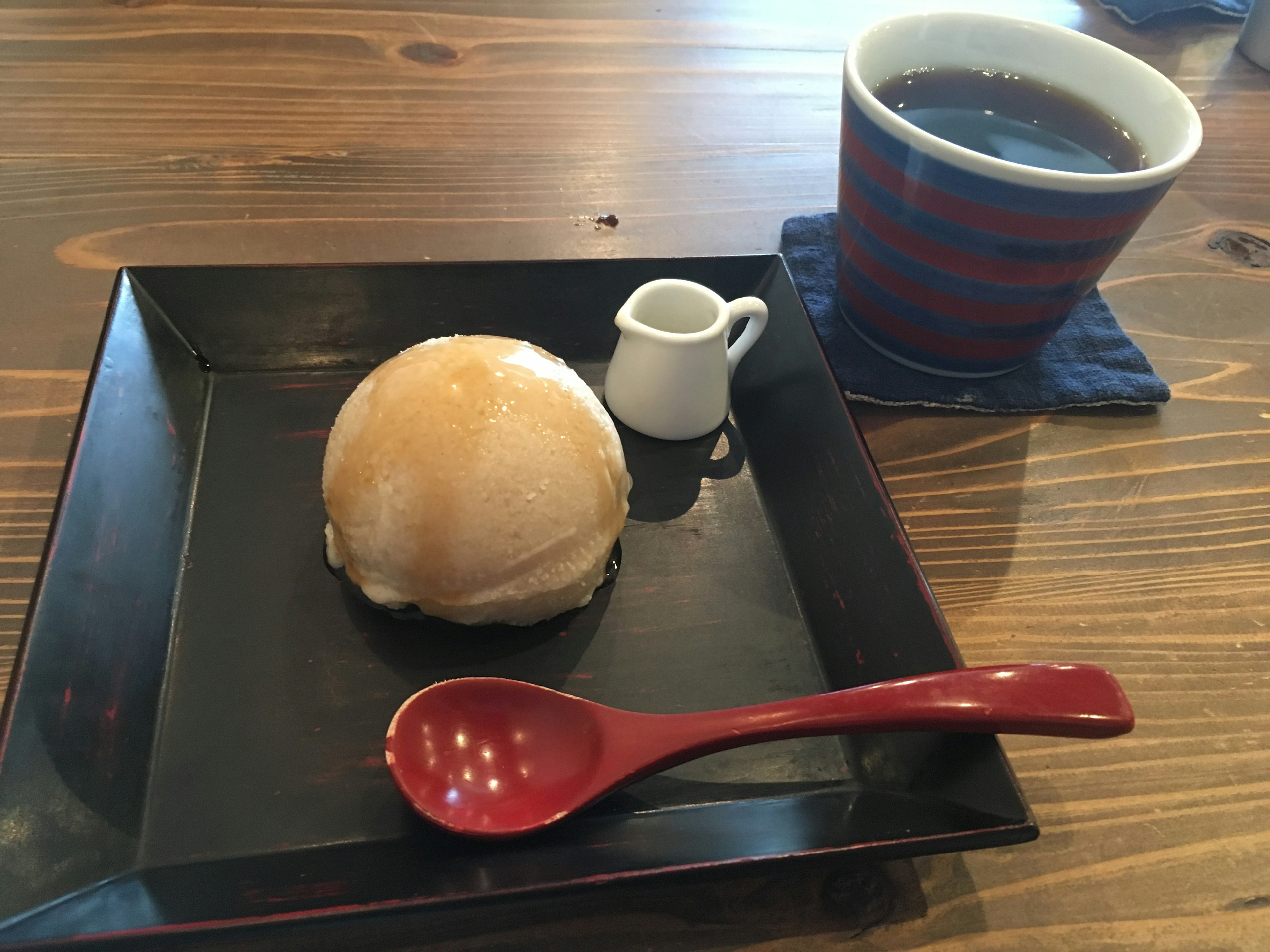 Un piatto nero con un panino bianco una piccola brocca di latte un cucchiaio rosso e una tazza di caffè a righe blu