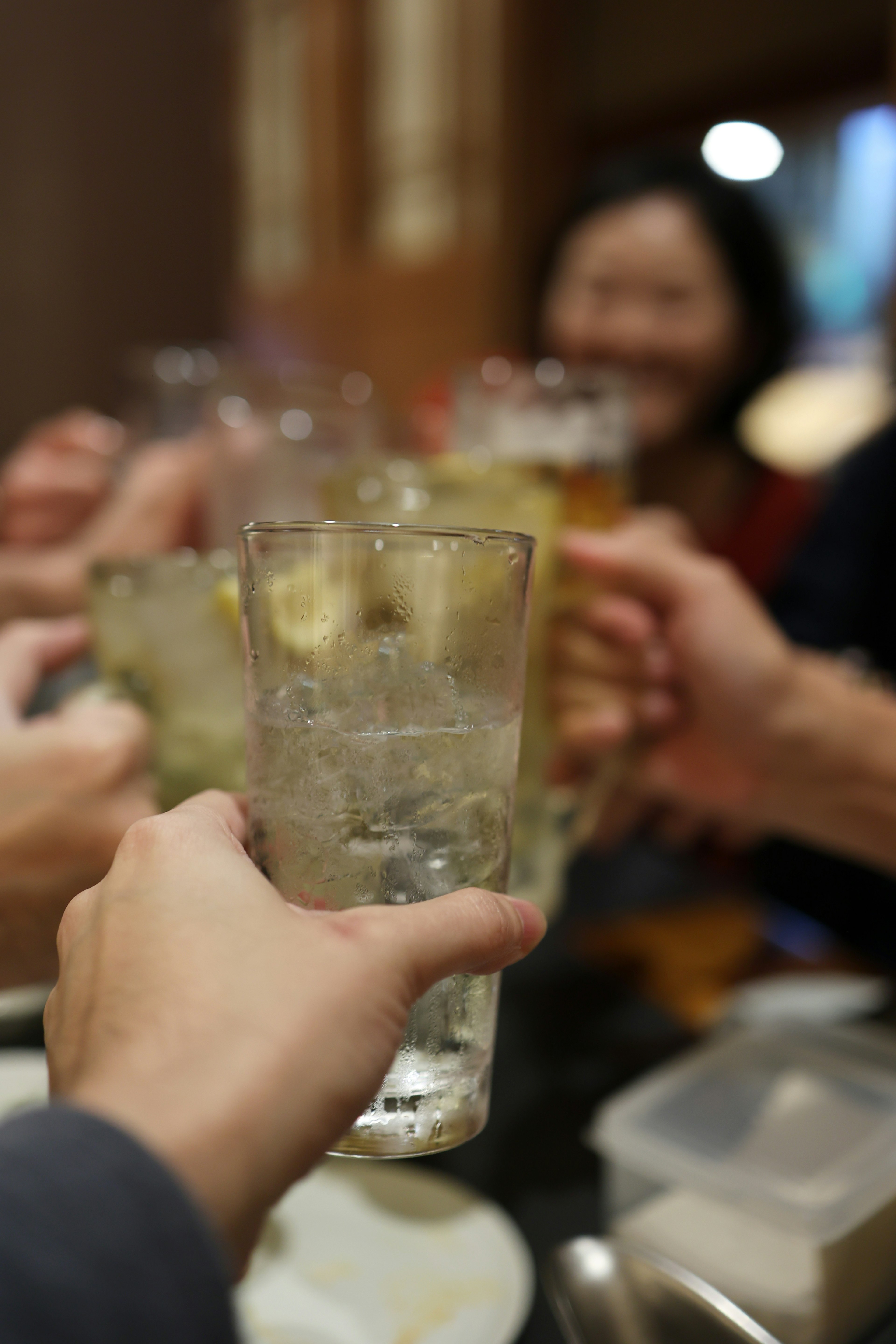 Manos sosteniendo vasos en un brindis con rostros sonrientes al fondo