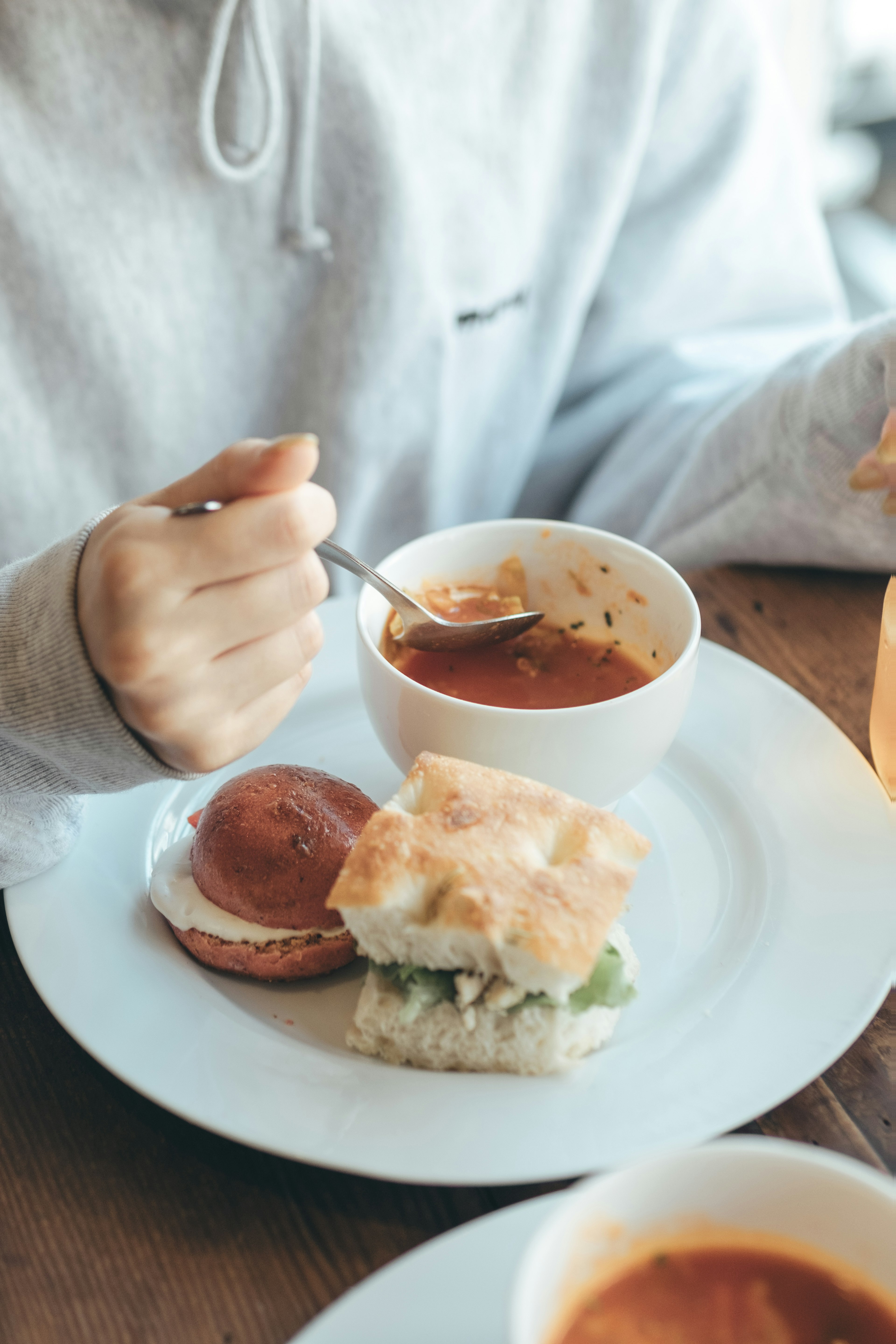 A hand in a sweatshirt holding a spoon enjoying soup with a sandwich on the plate