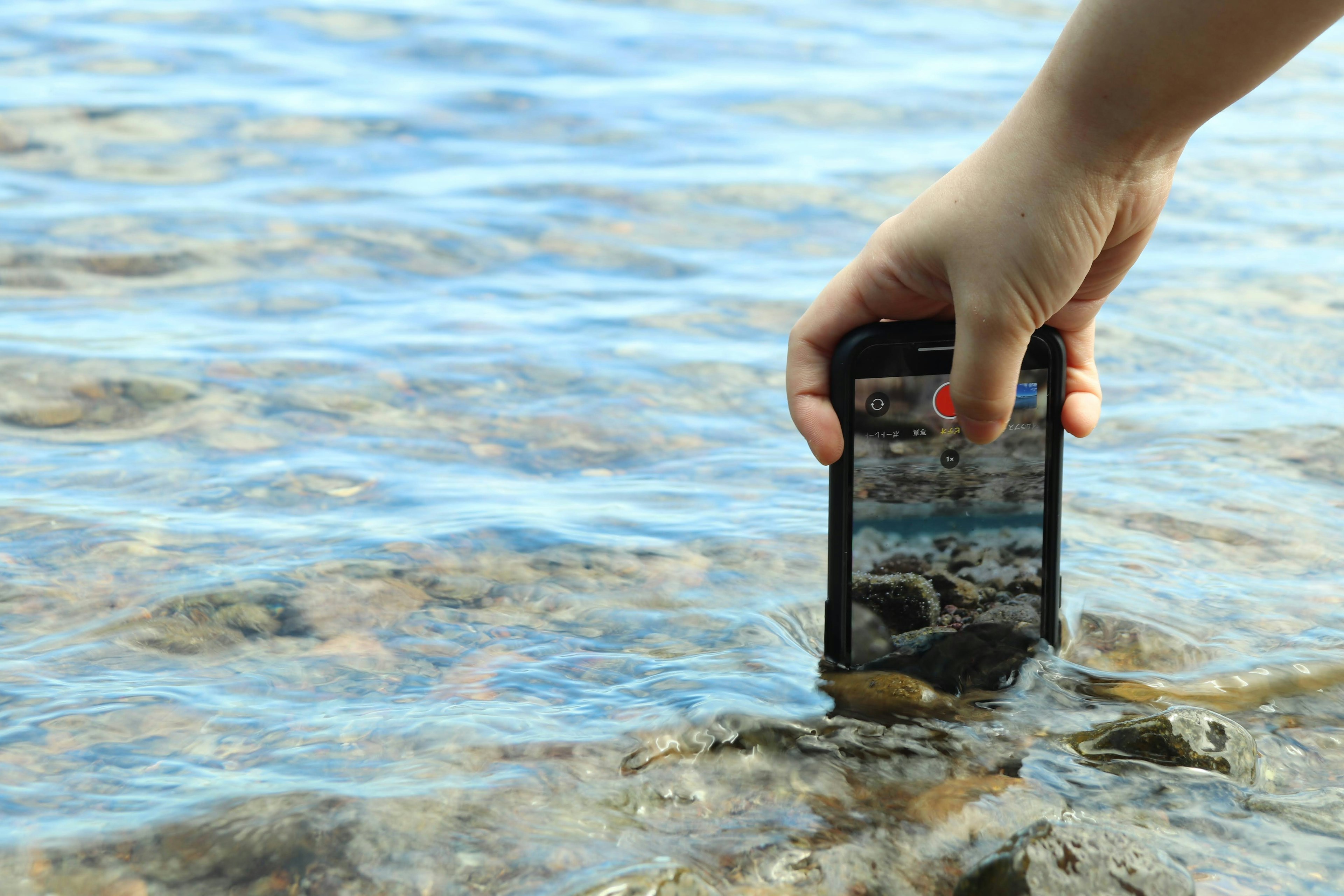 Hand holding a smartphone above water surface with reflections and rocks