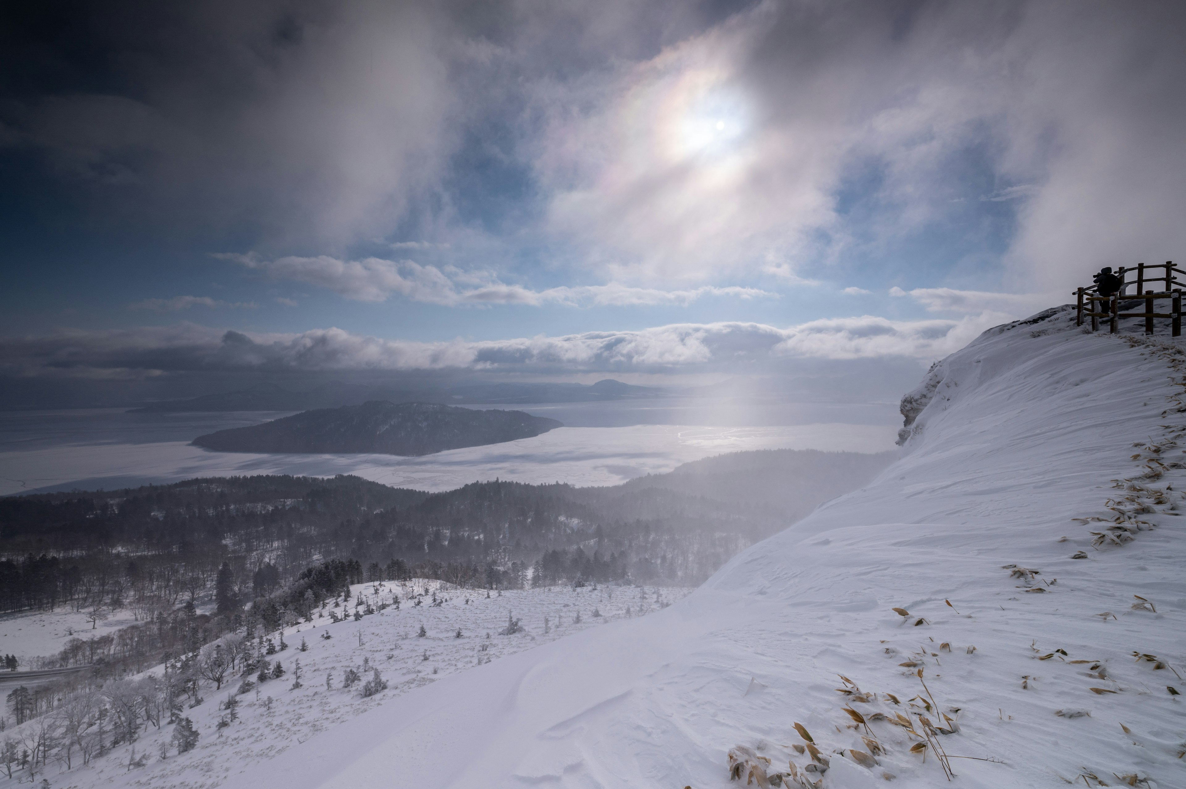 雪に覆われた山の頂上からの風景 雲がかかった空と太陽の光が反射する湖