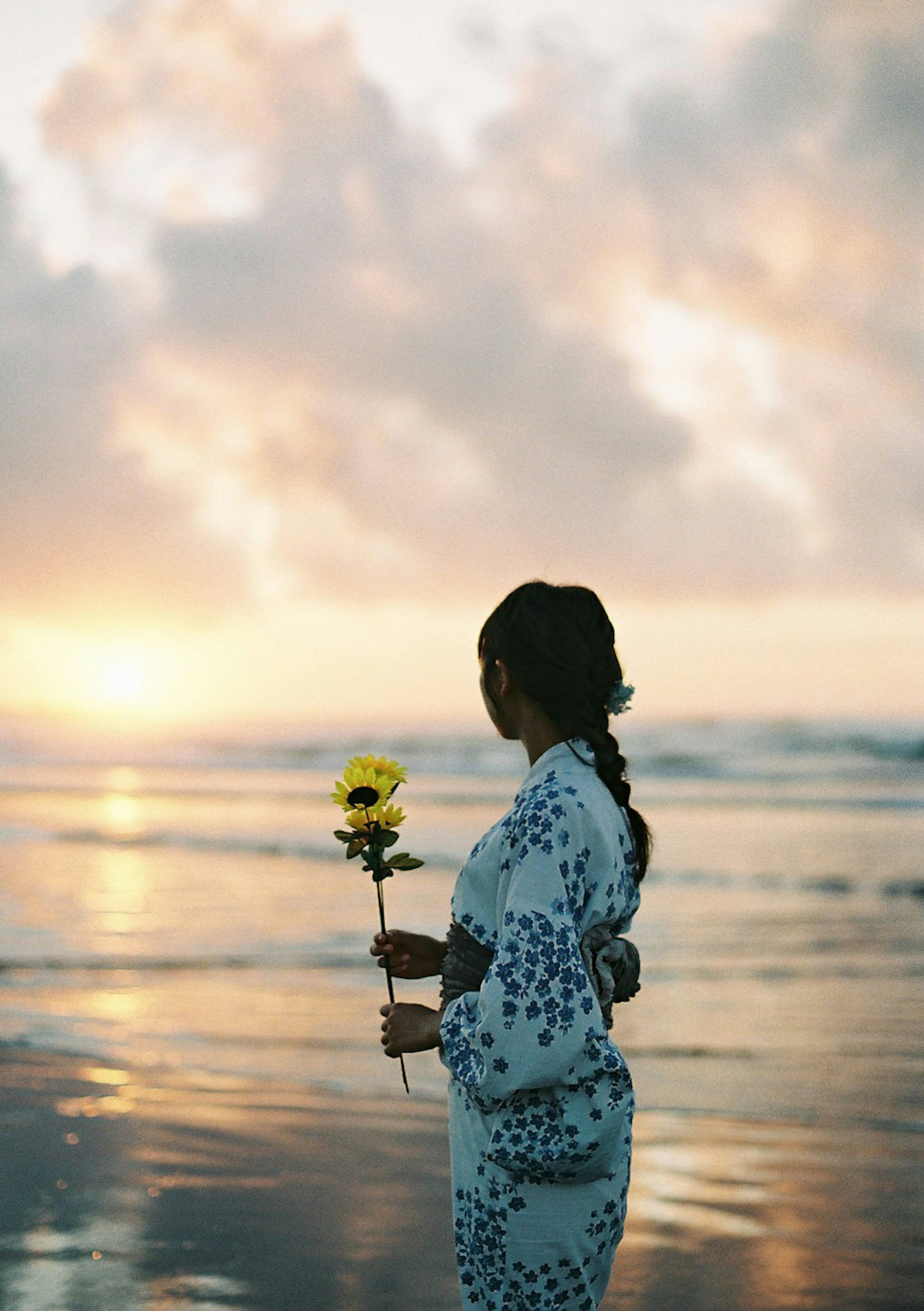 ビーチに立つ女性が花を持ちながら夕日を見つめている