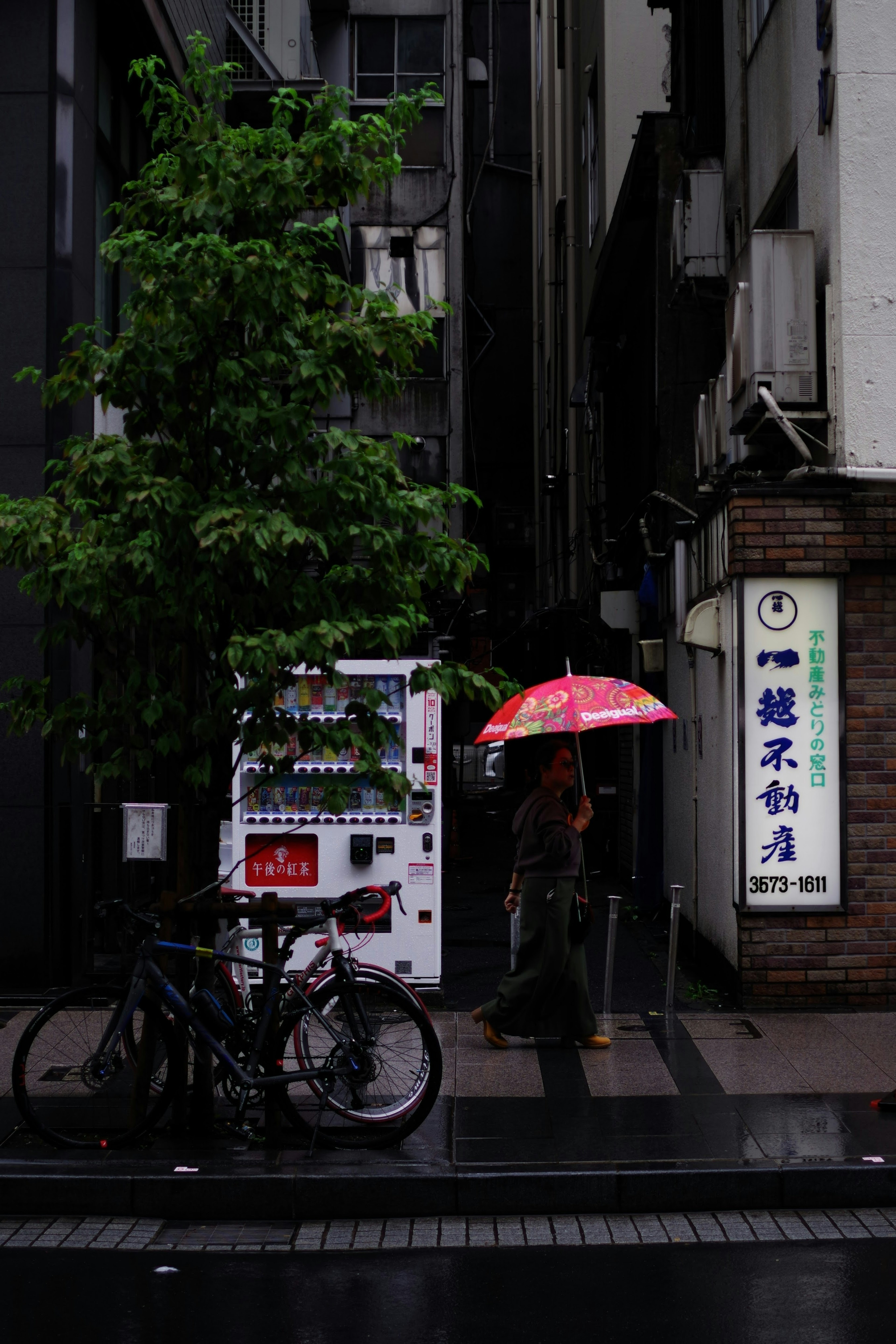 雨の日の街角にある自動販売機と赤い傘を持った人物