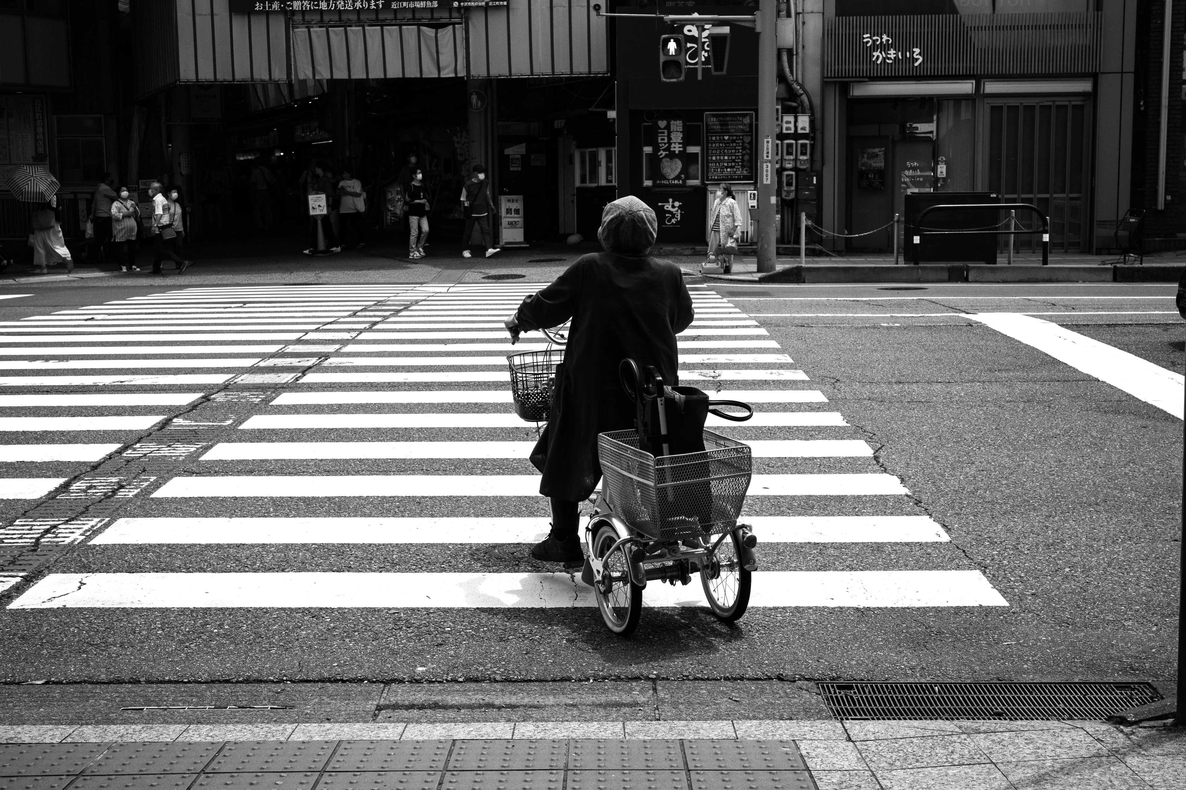 Black and white photo of an elderly person crossing a crosswalk with a shopping cart