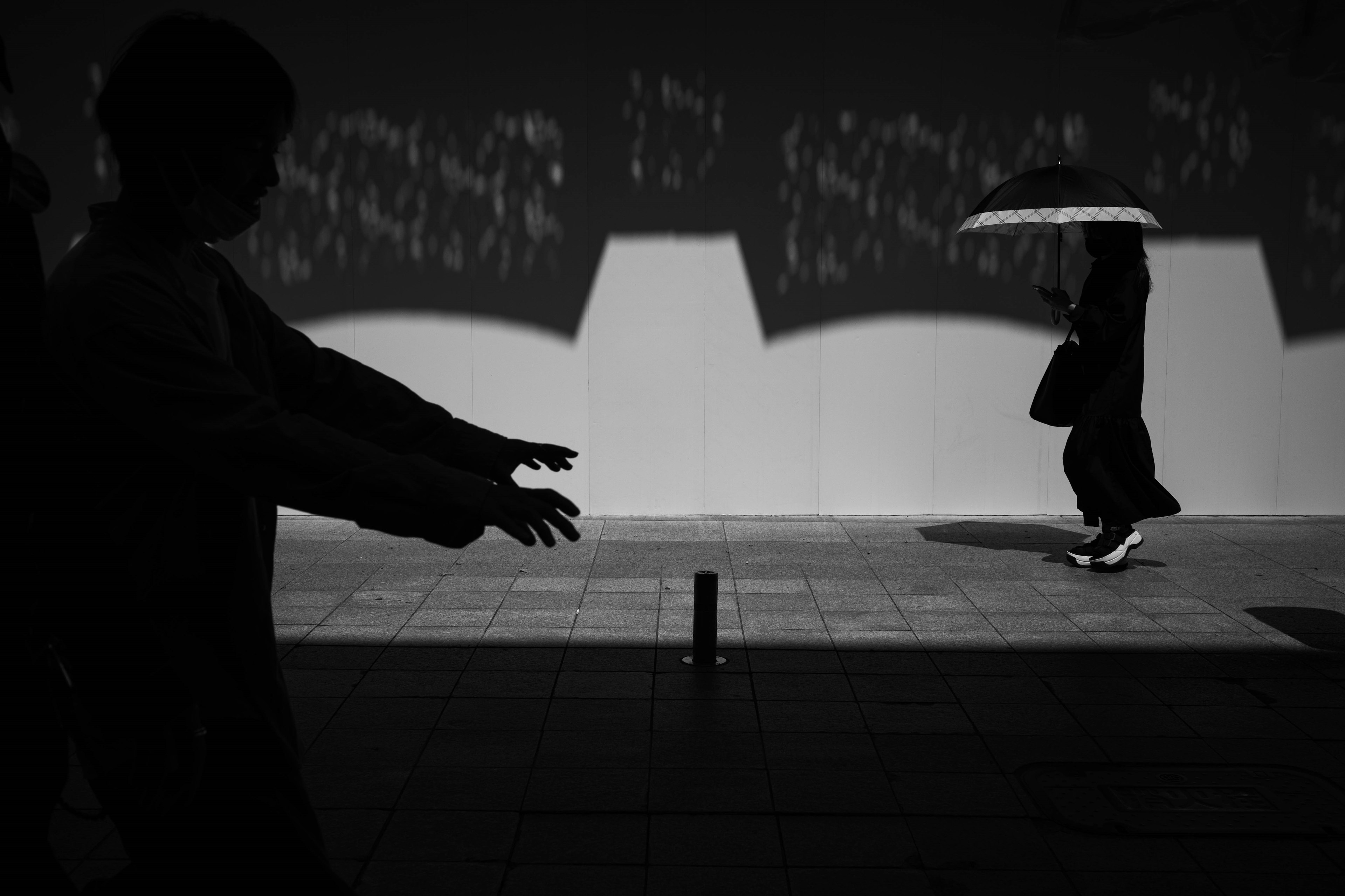 A street scene depicted in black and white contrast featuring shadows of people and a woman walking with an umbrella