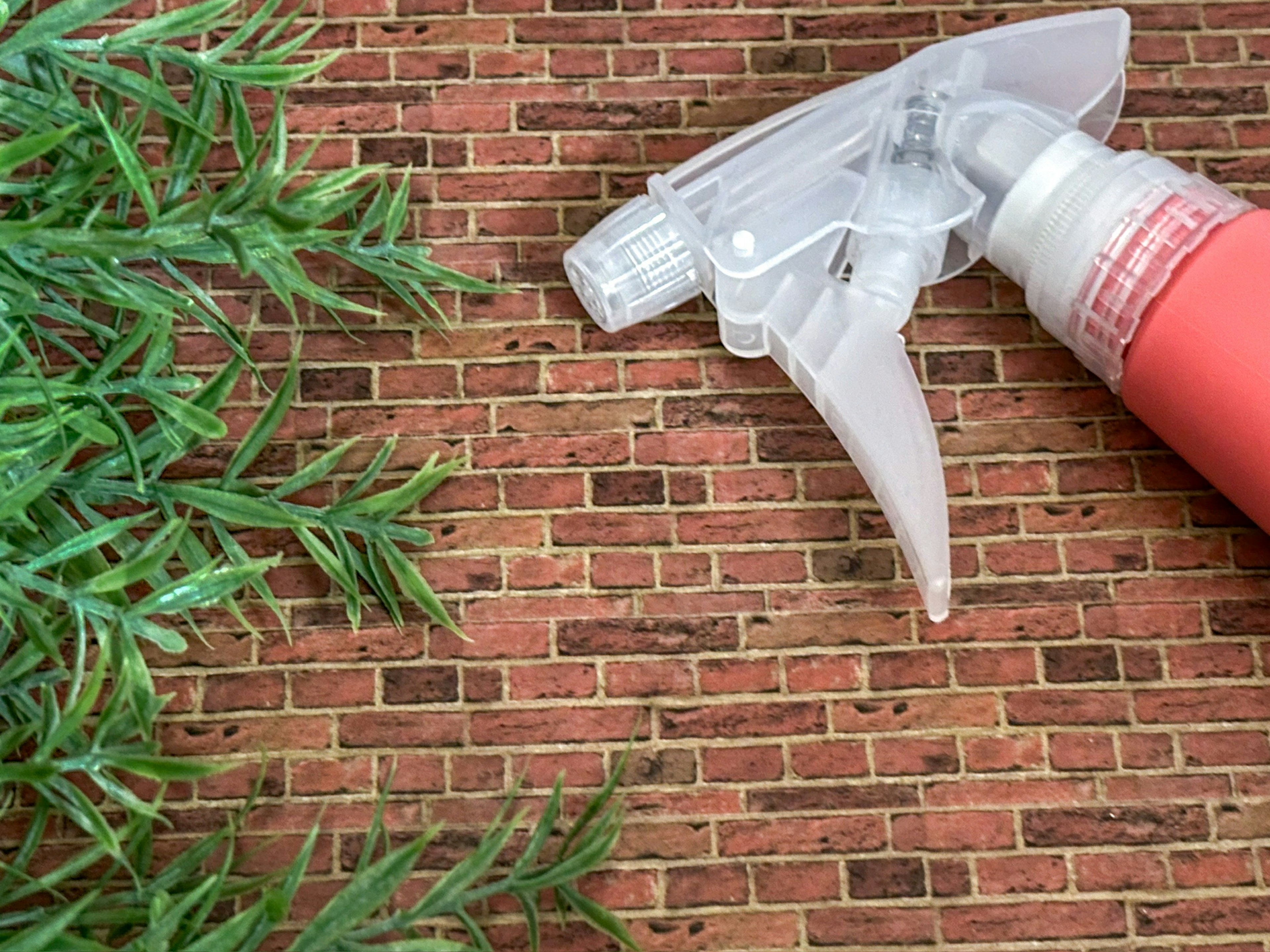 Image featuring a red spray bottle and green plant against a brick background