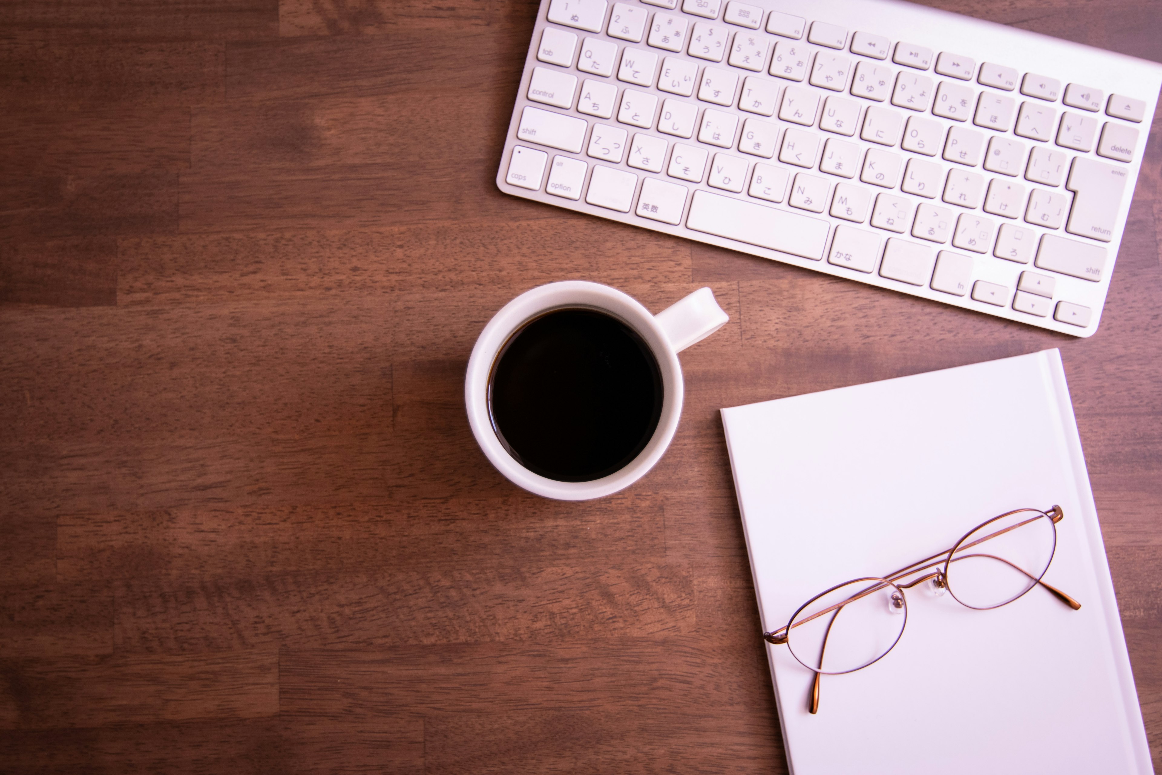 Teclado blanco y taza de café sobre una mesa de madera cuaderno y gafas al lado