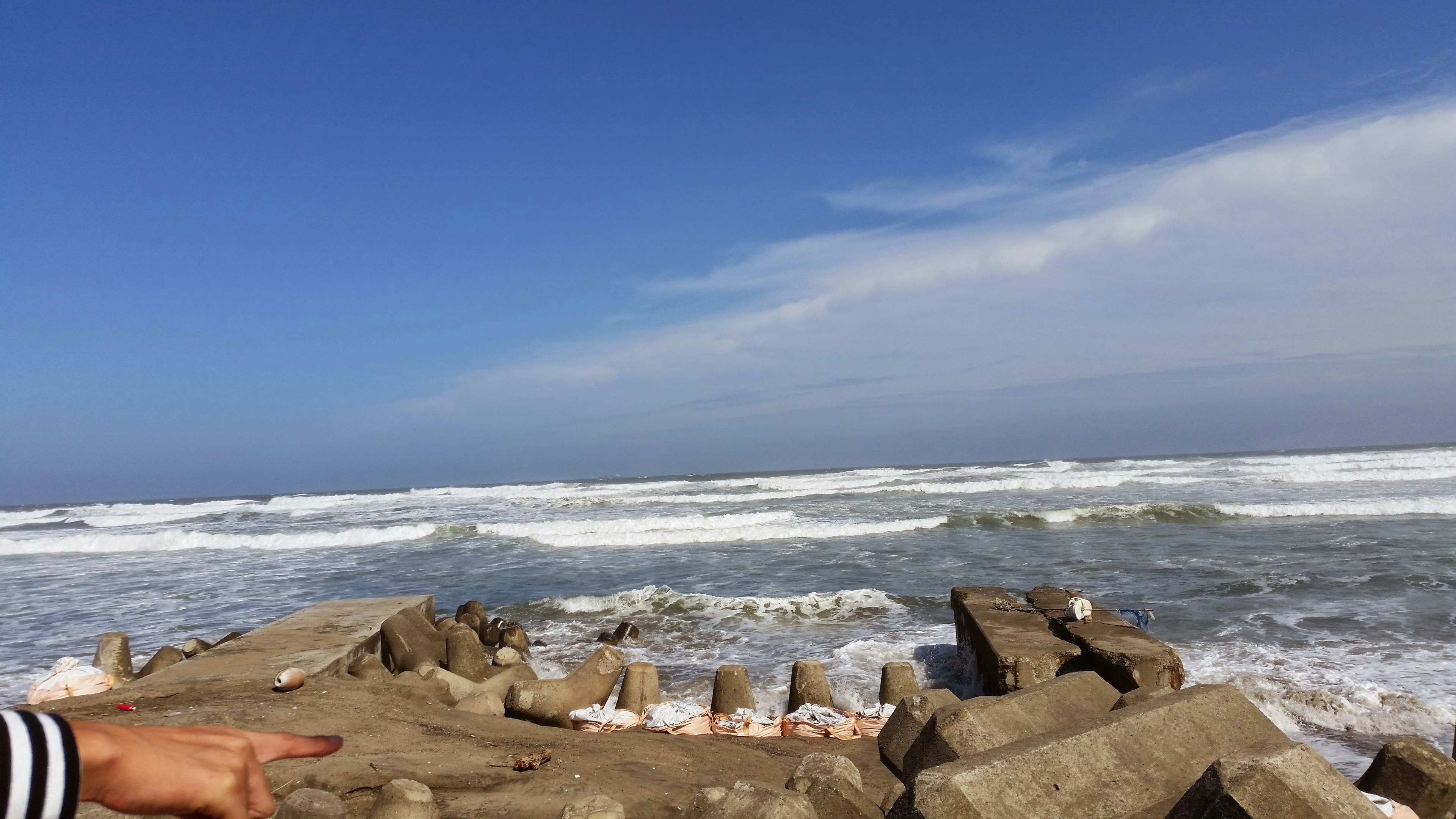 Scène côtière avec ciel bleu et vagues s'écrasant Formations rocheuses au premier plan avec une personne pointant