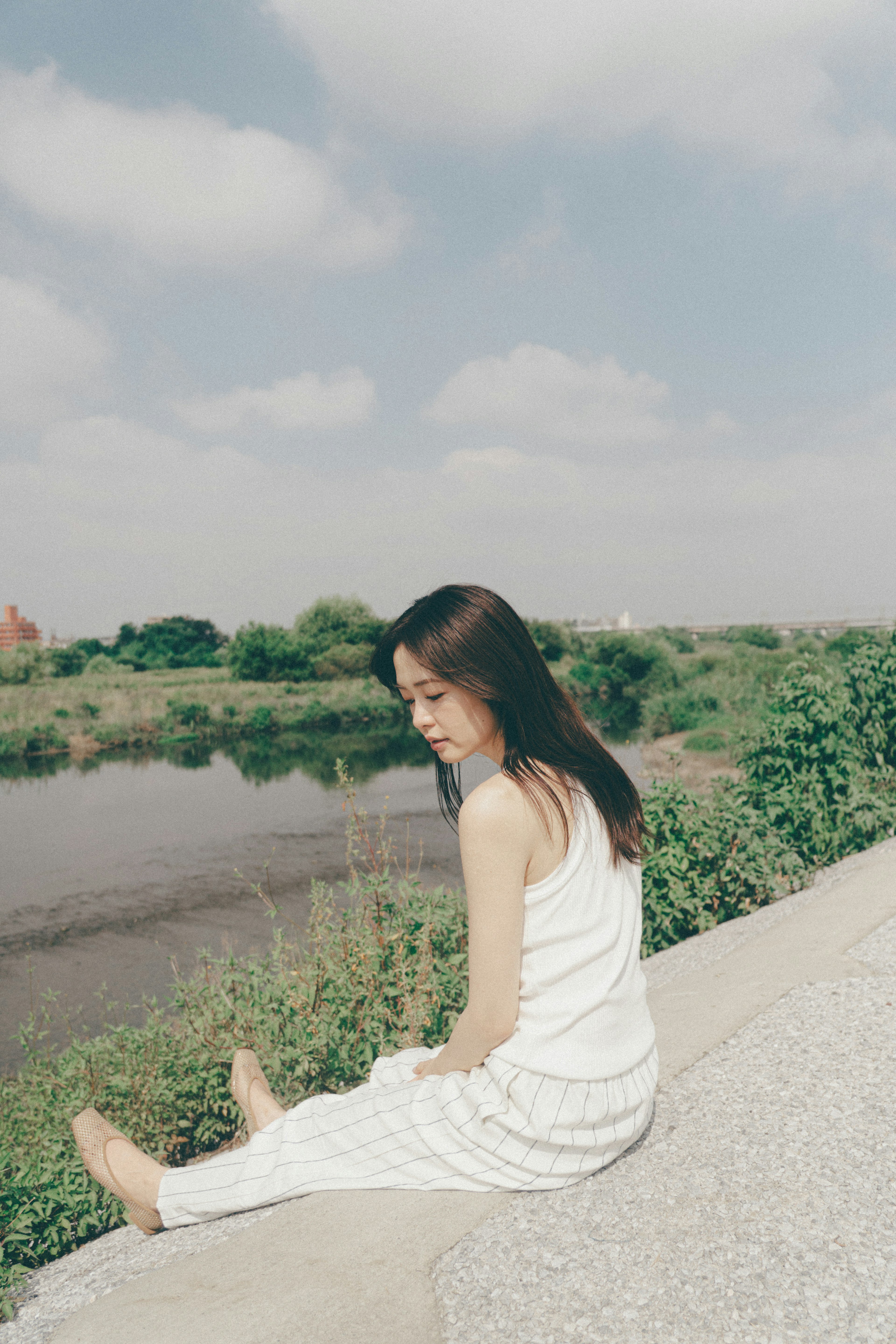 Woman sitting by the river wearing white clothes green grass and blue sky