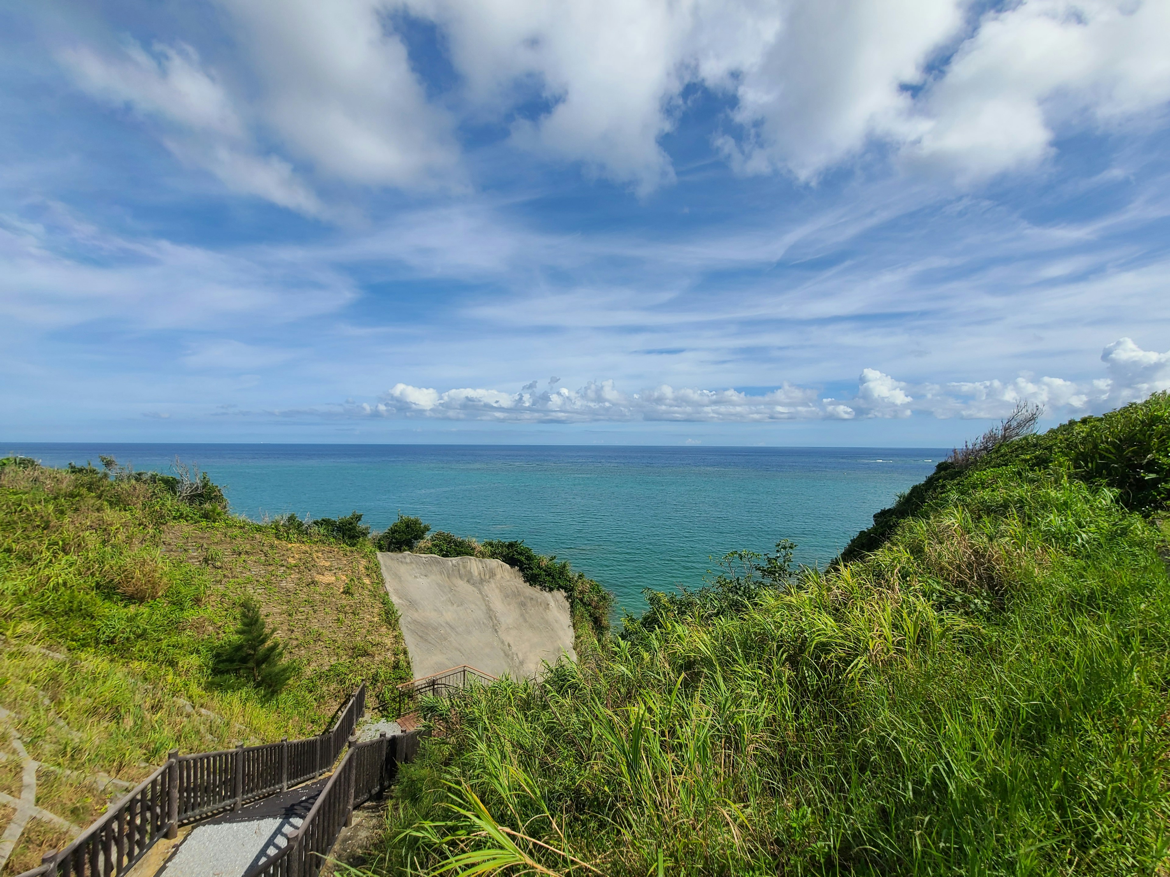 Pemandangan laut biru dan langit dengan bukit hijau subur dan tangga