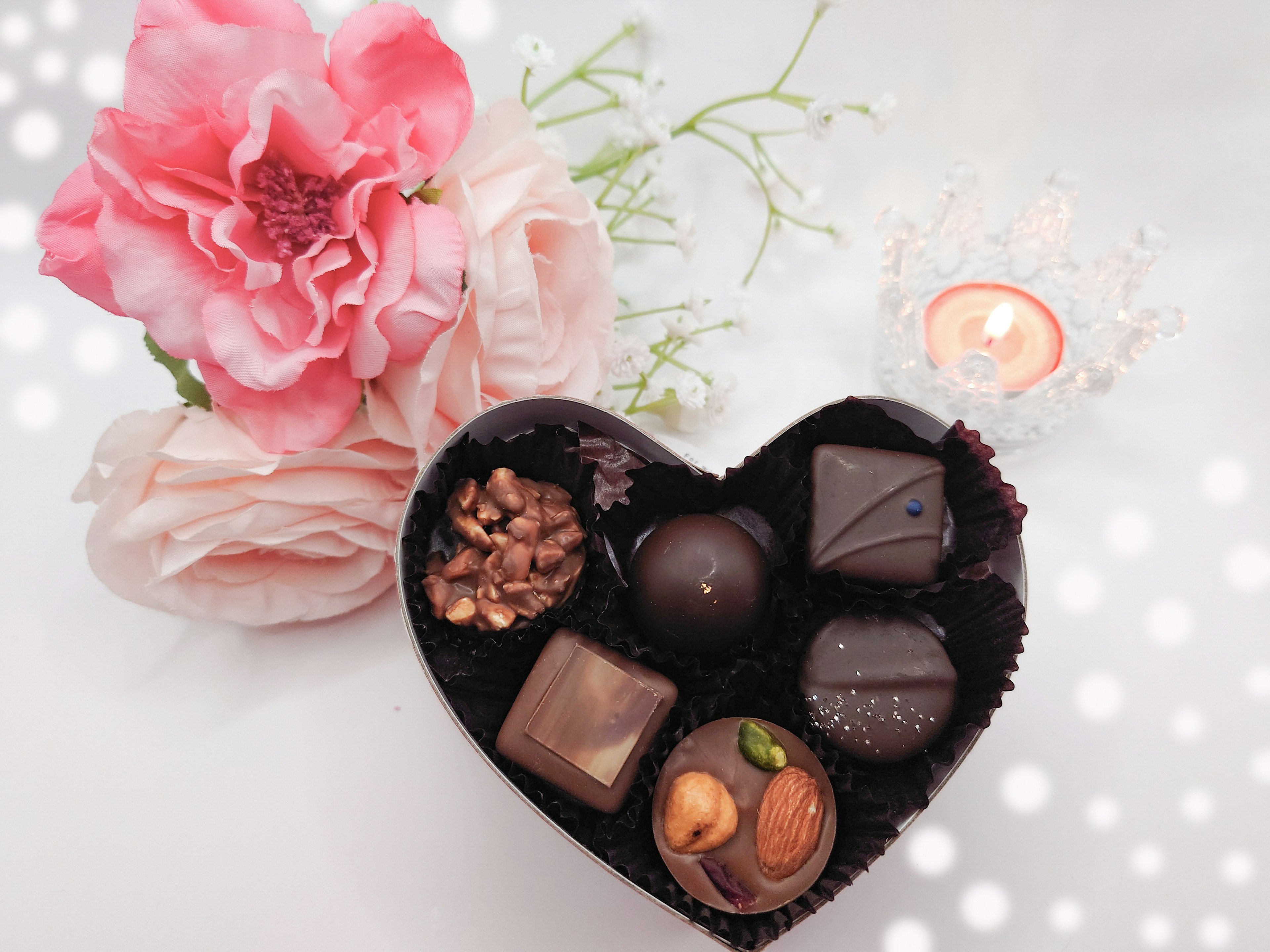 Una caja en forma de corazón llena de chocolates variados junto a una flor rosa y una vela