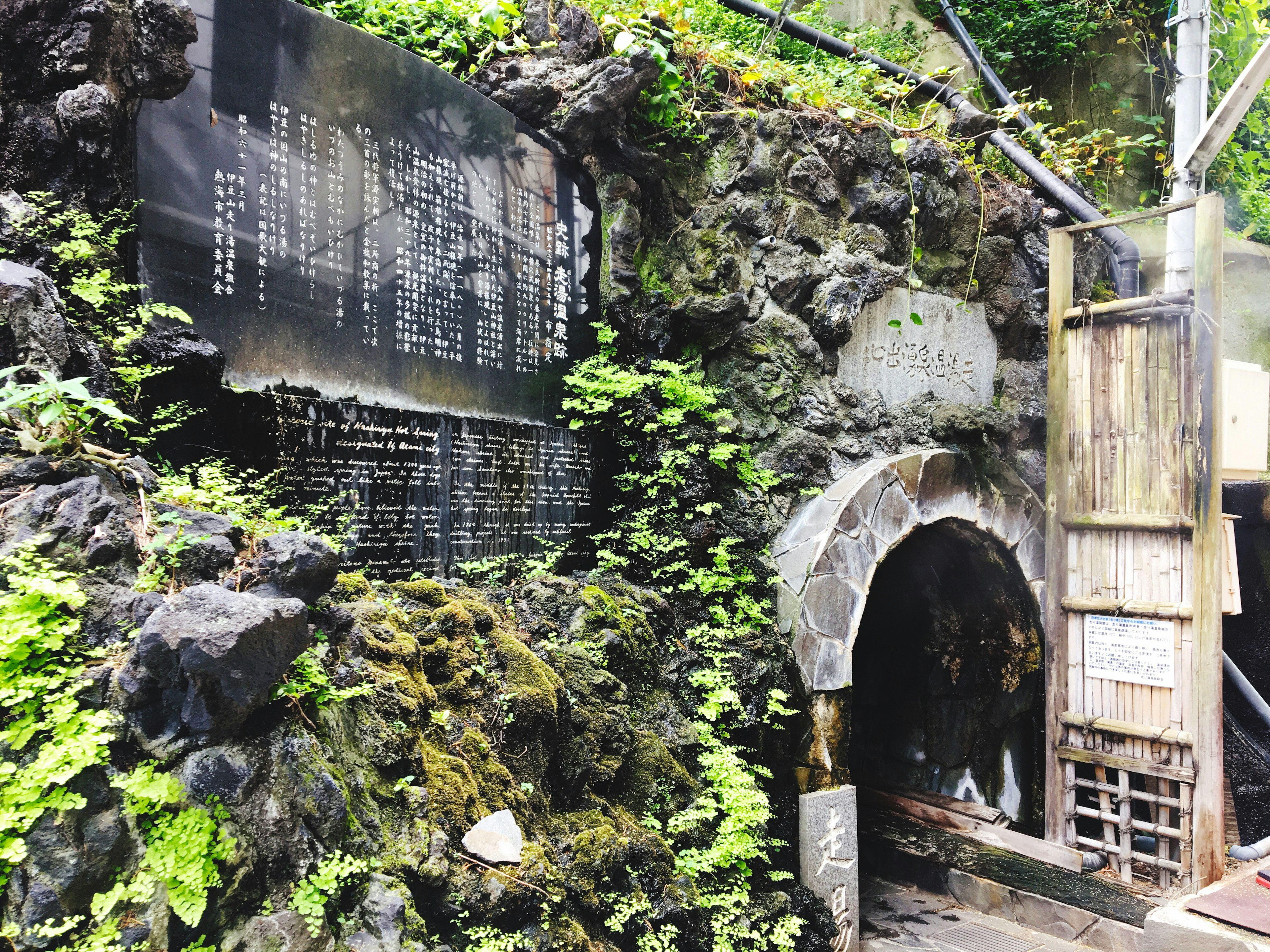 岩や苔に覆われた温泉の入口と説明看板がある風景