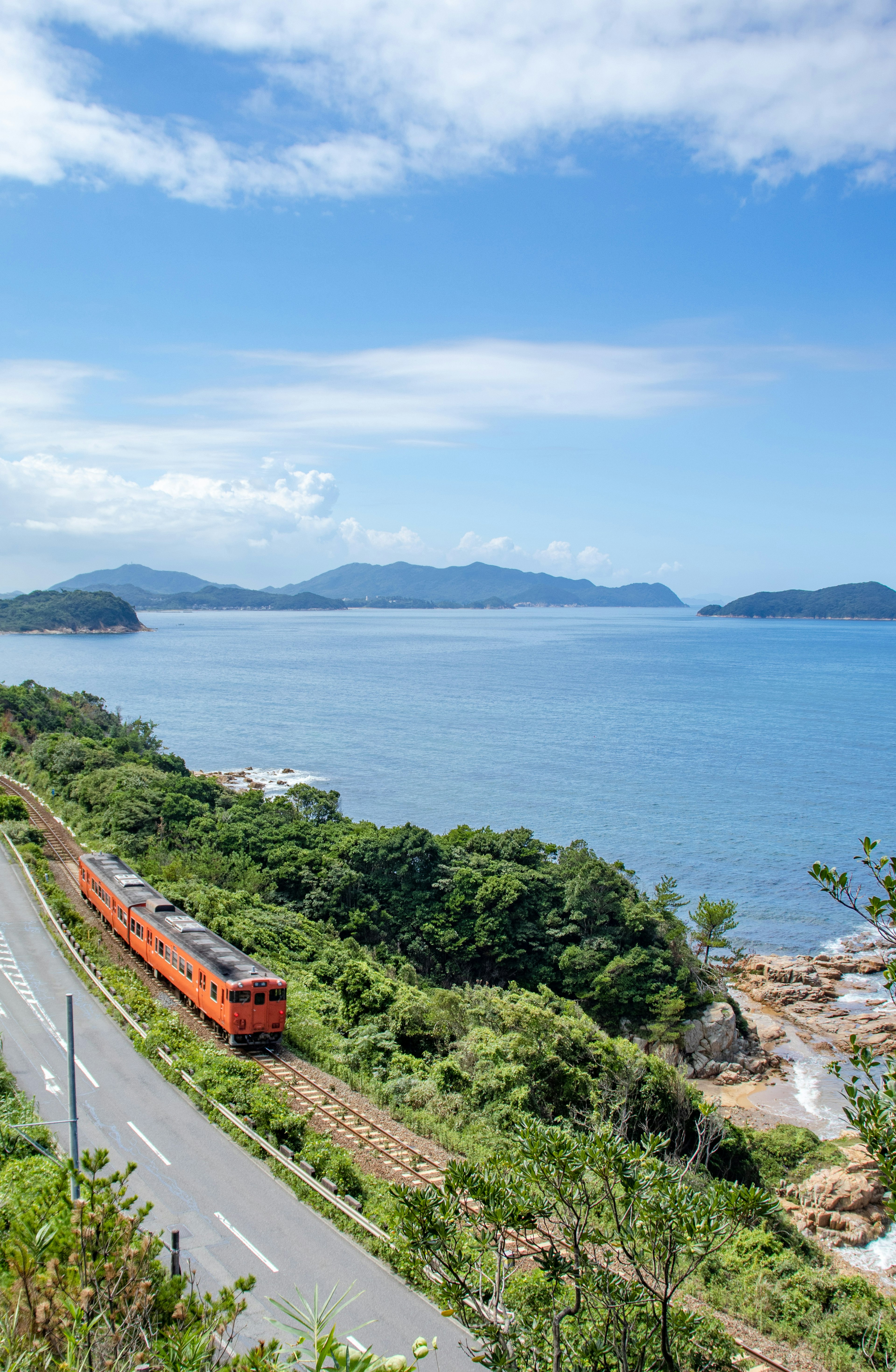 Kereta api oranye yang melaju di sepanjang laut biru dan bukit hijau