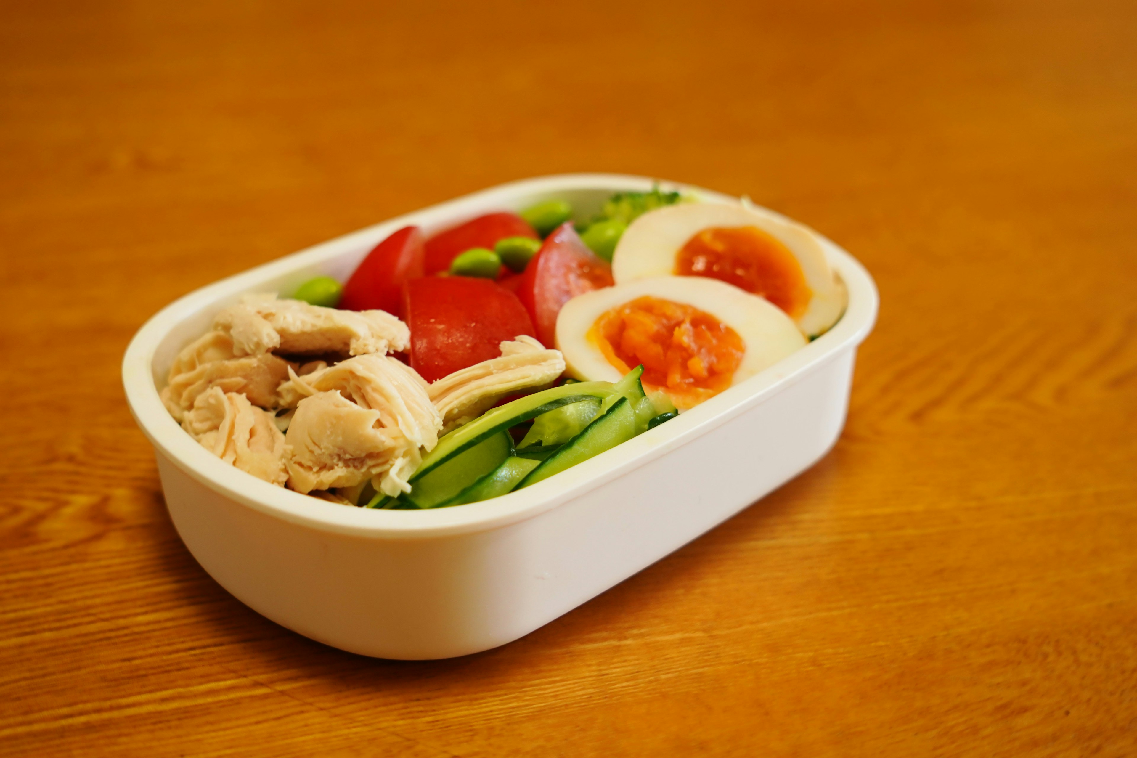 A white bento box filled with chicken, tomatoes, green onions, and soft-boiled eggs