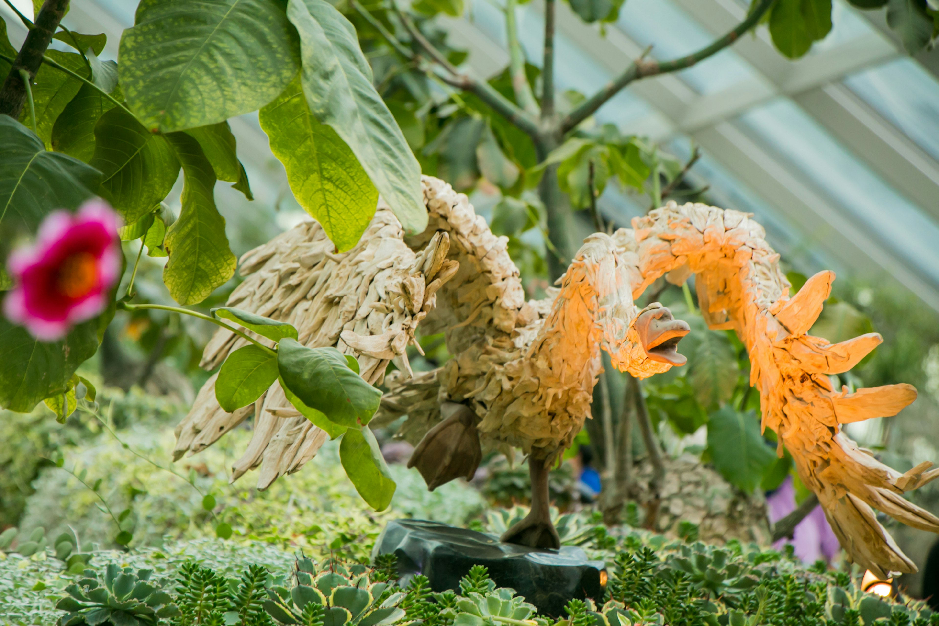 緑豊かな温室の中で、色とりどりの植物に囲まれたアート作品の鶴が見える