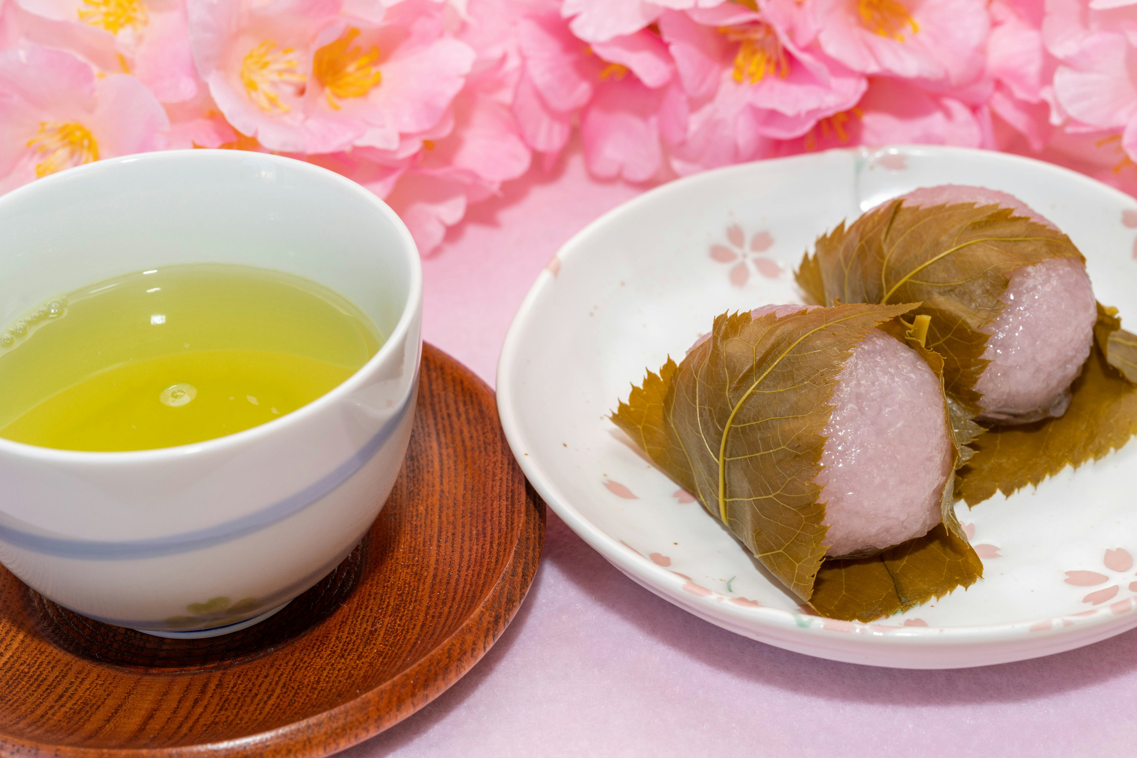 A bowl of green tea next to sakura mochi on a plate with pink cherry blossoms in the background