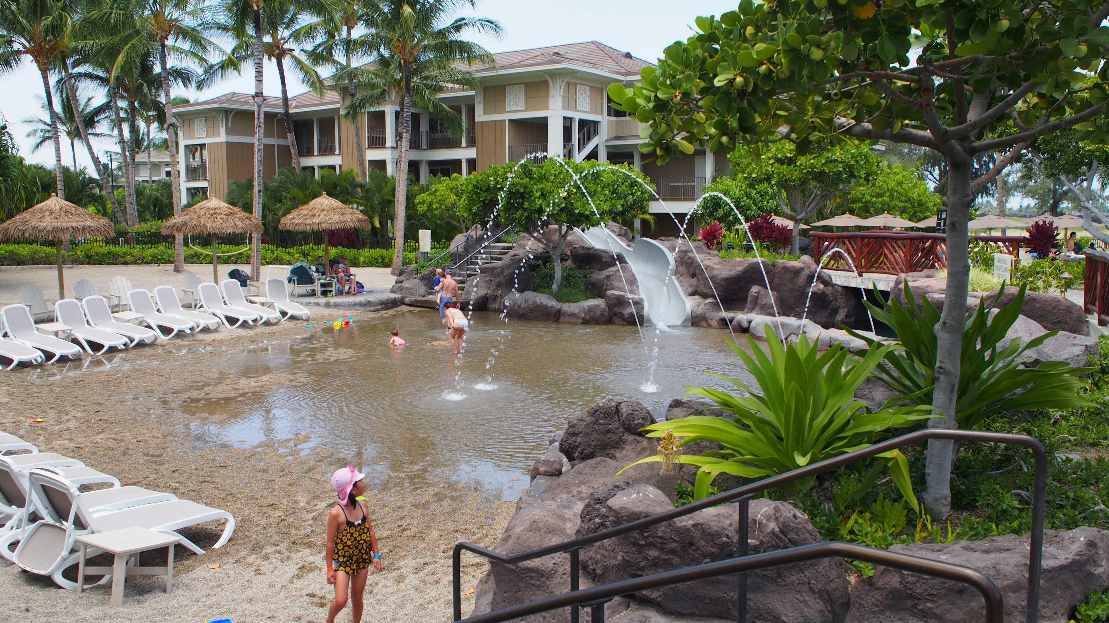 Niños jugando en un área de piscina de resort con una cascada y vegetación exuberante