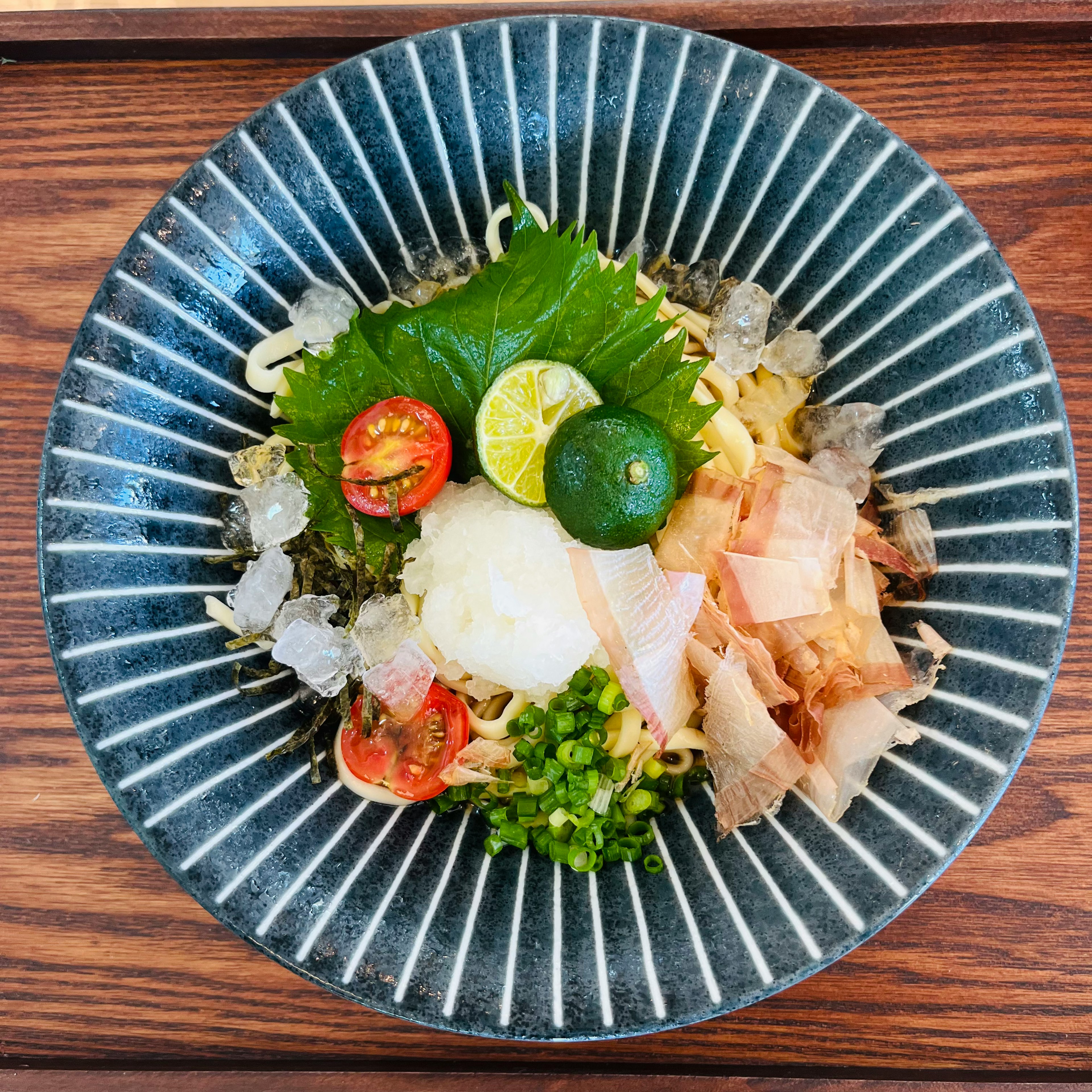 Schön präsentierte japanische kalte Nudeln in einer blauen gestreiften Schüssel mit Tomaten, Frühlingszwiebeln, geriebenem Rettich, Shiso-Blättern und Zitrusfrüchten