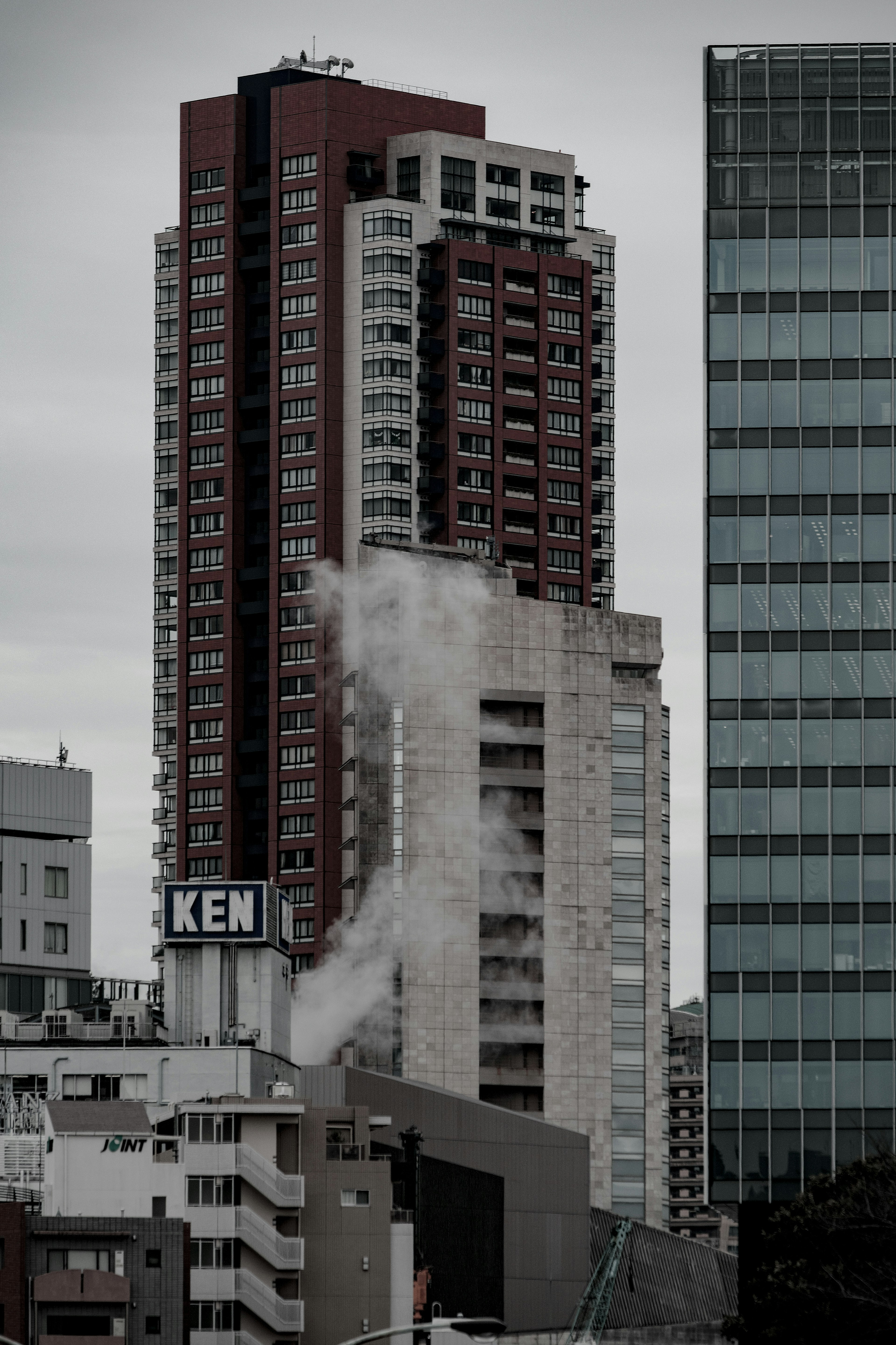 A tall red building amidst urban skyscrapers with smoke rising