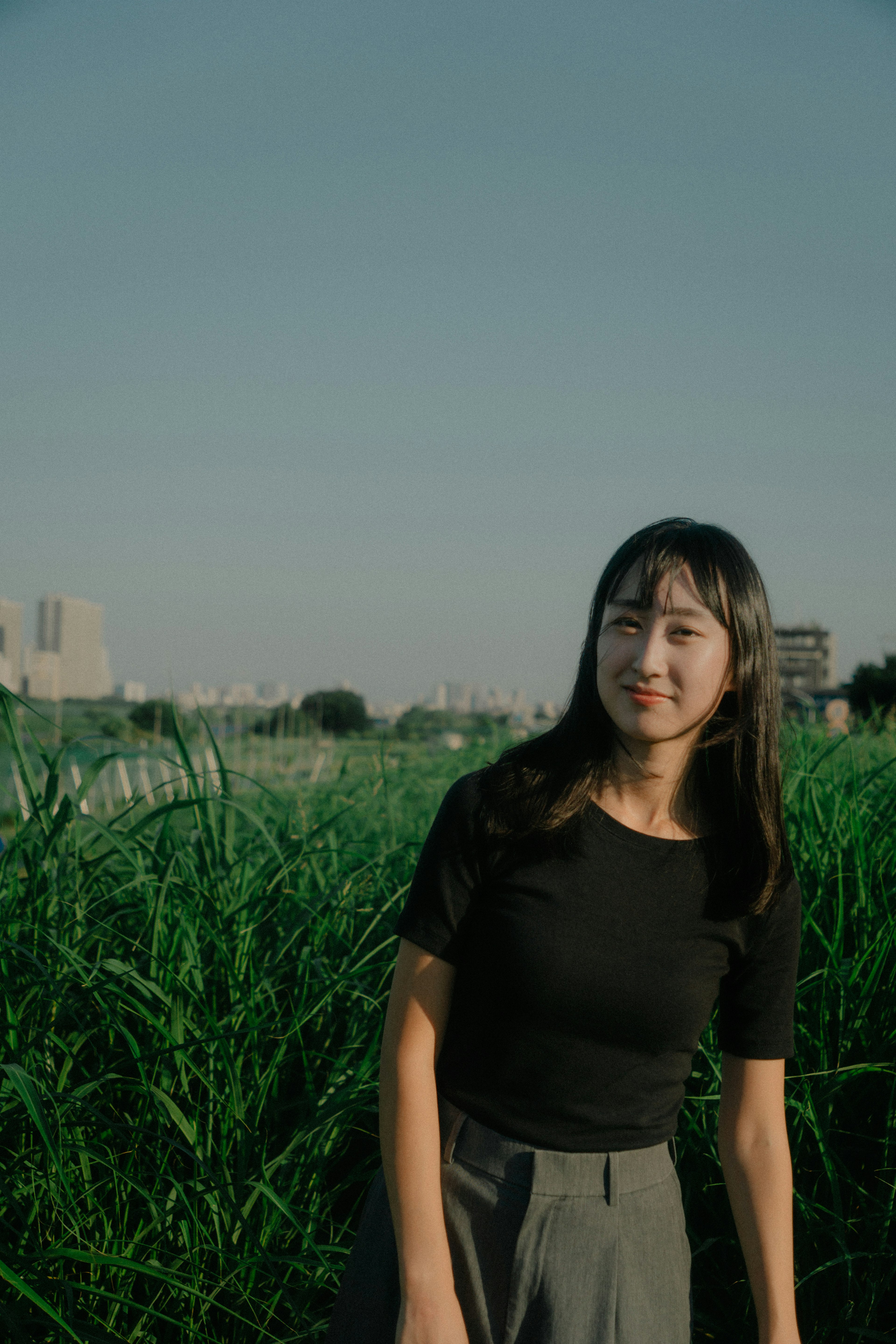 草原に立つ女性のポートレート　黒いTシャツとグレーのパンツを着用　背景には都会の風景が広がる