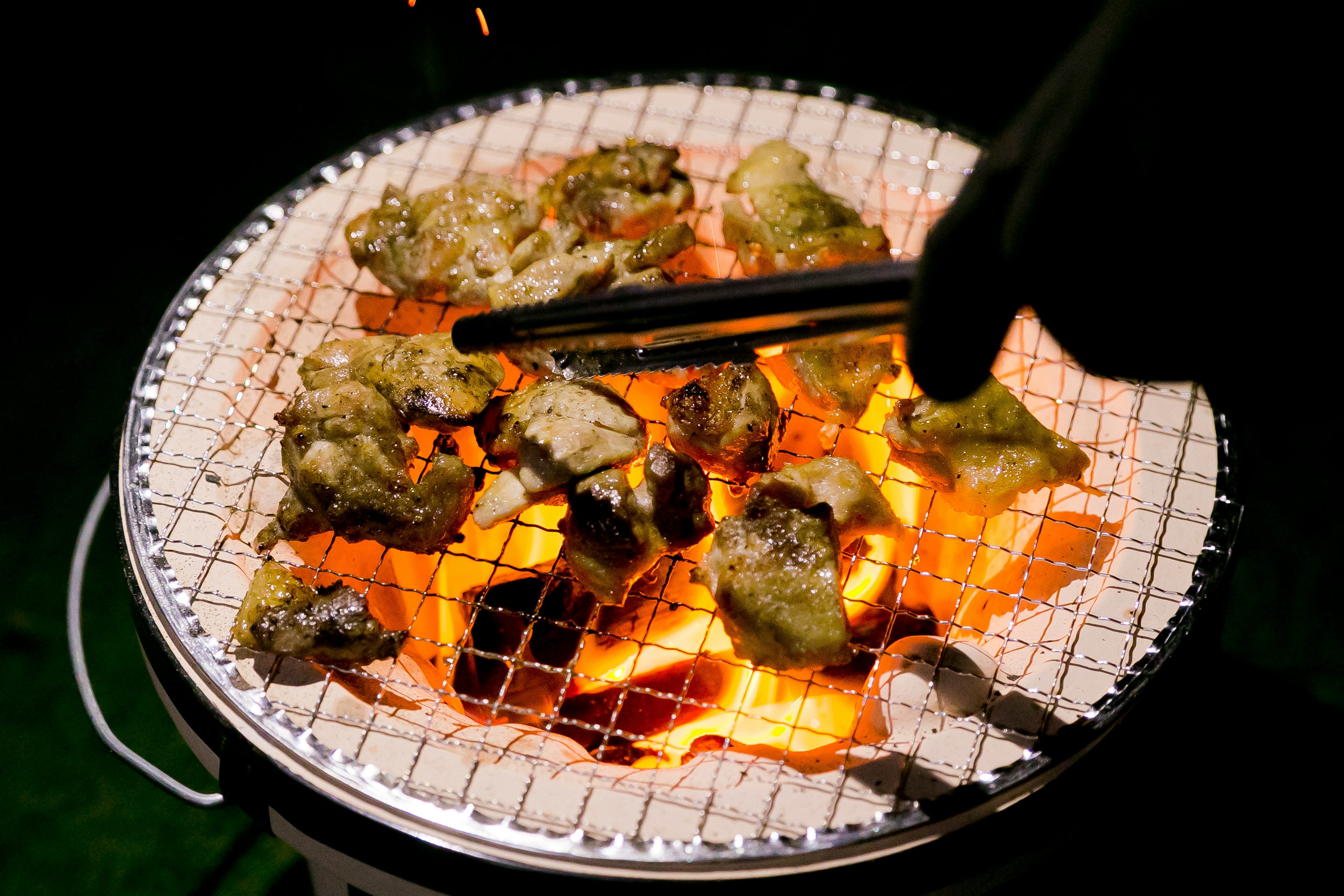 Grilled meat pieces on a charcoal barbecue with a hand holding tongs