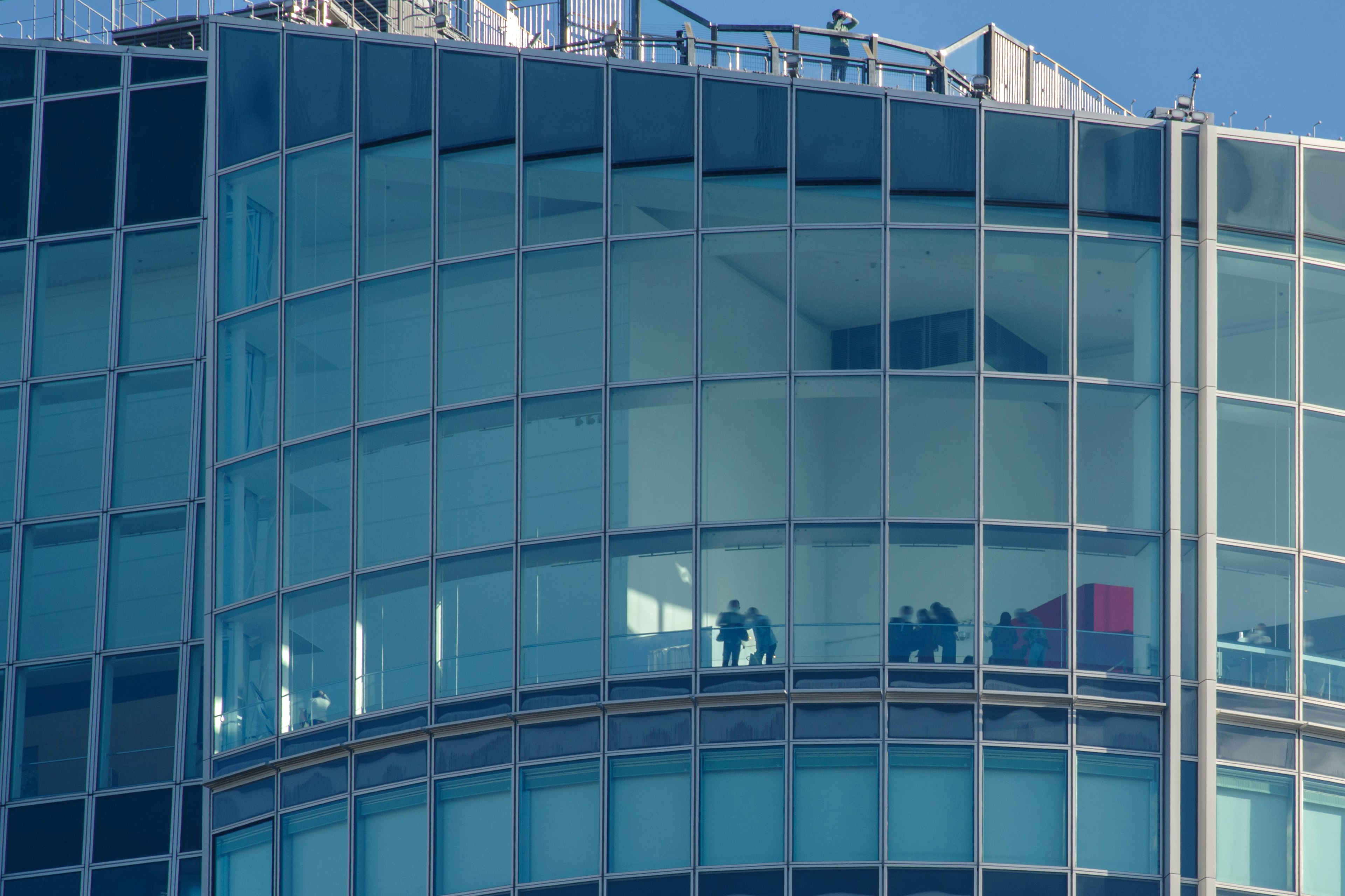 People visible through the glass windows of a skyscraper with workers on the rooftop