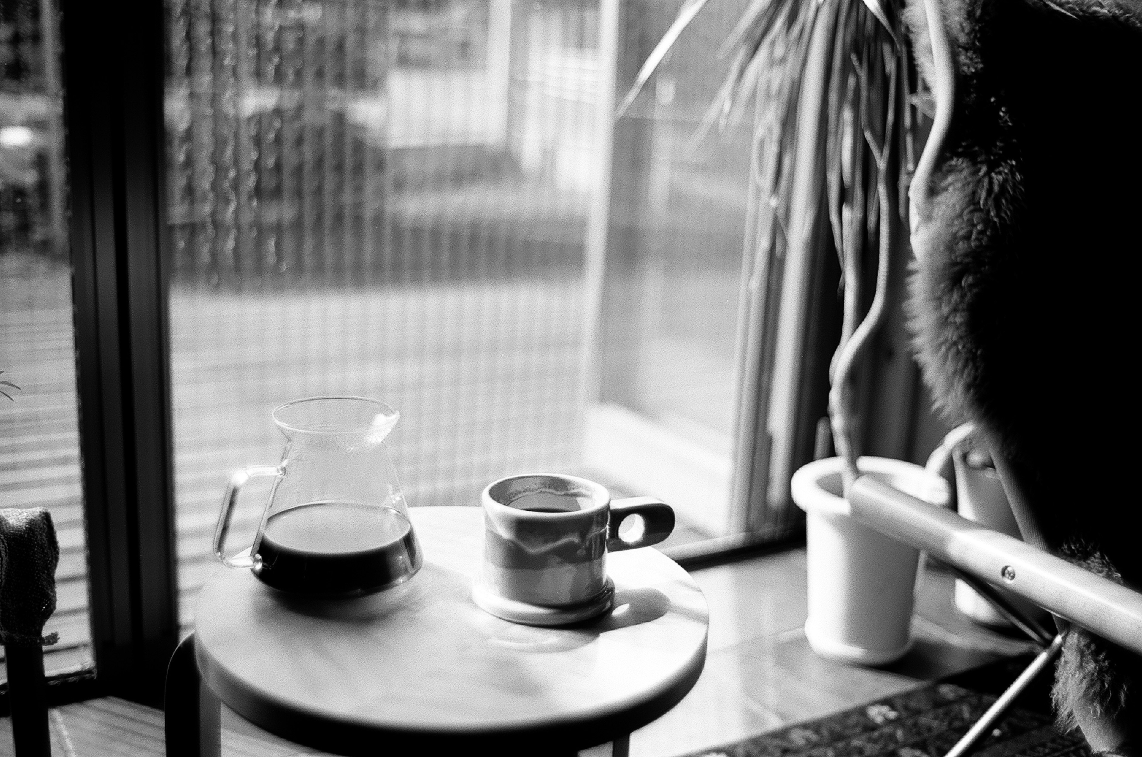 Schwarzweißbild eines Cafétisches mit einer Tasse Kaffee und einer Kanne Natürliches Licht, das durch ein Fenster strömt