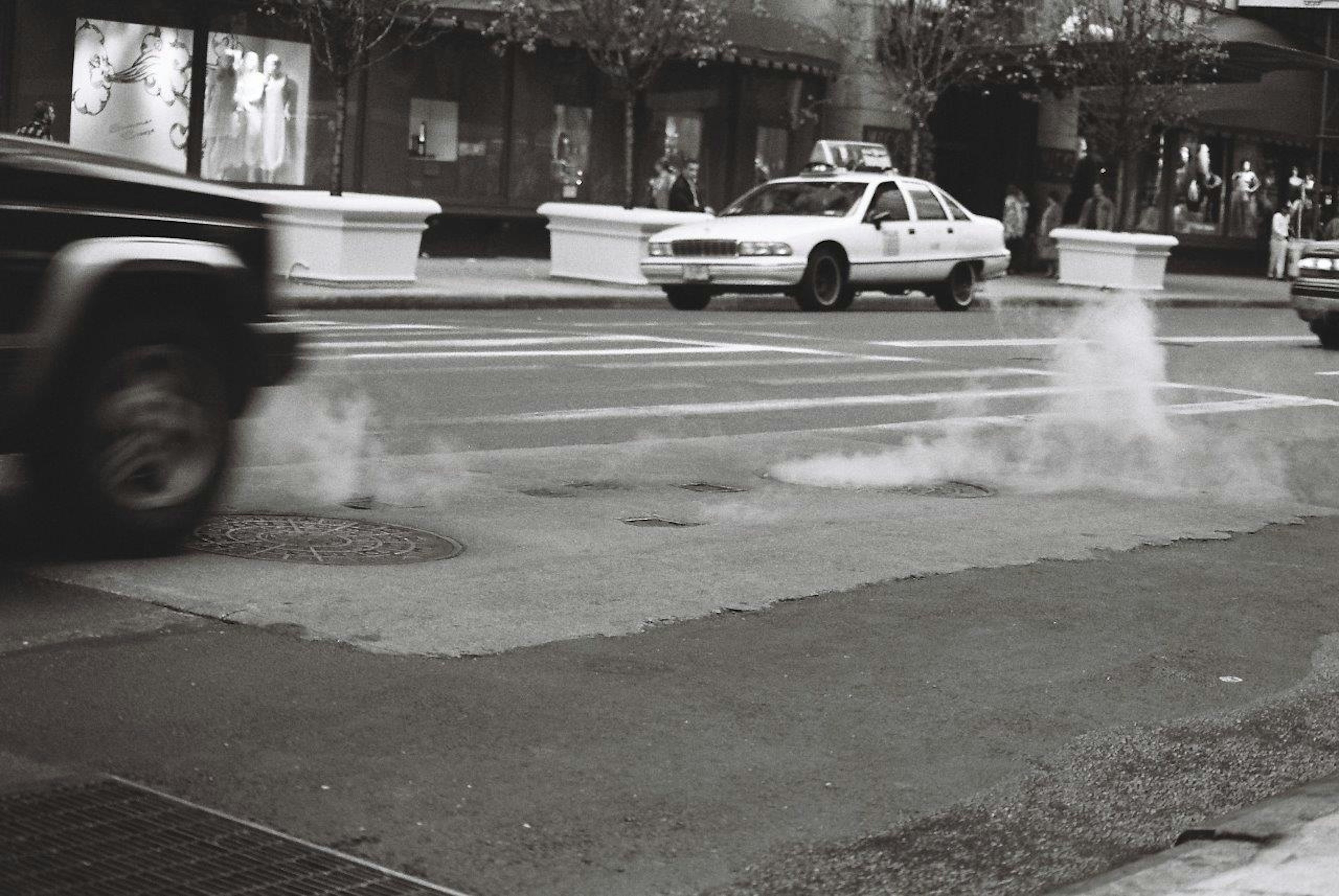 Black and white city scene with a taxi and a jeep passing through an intersection with steam rising