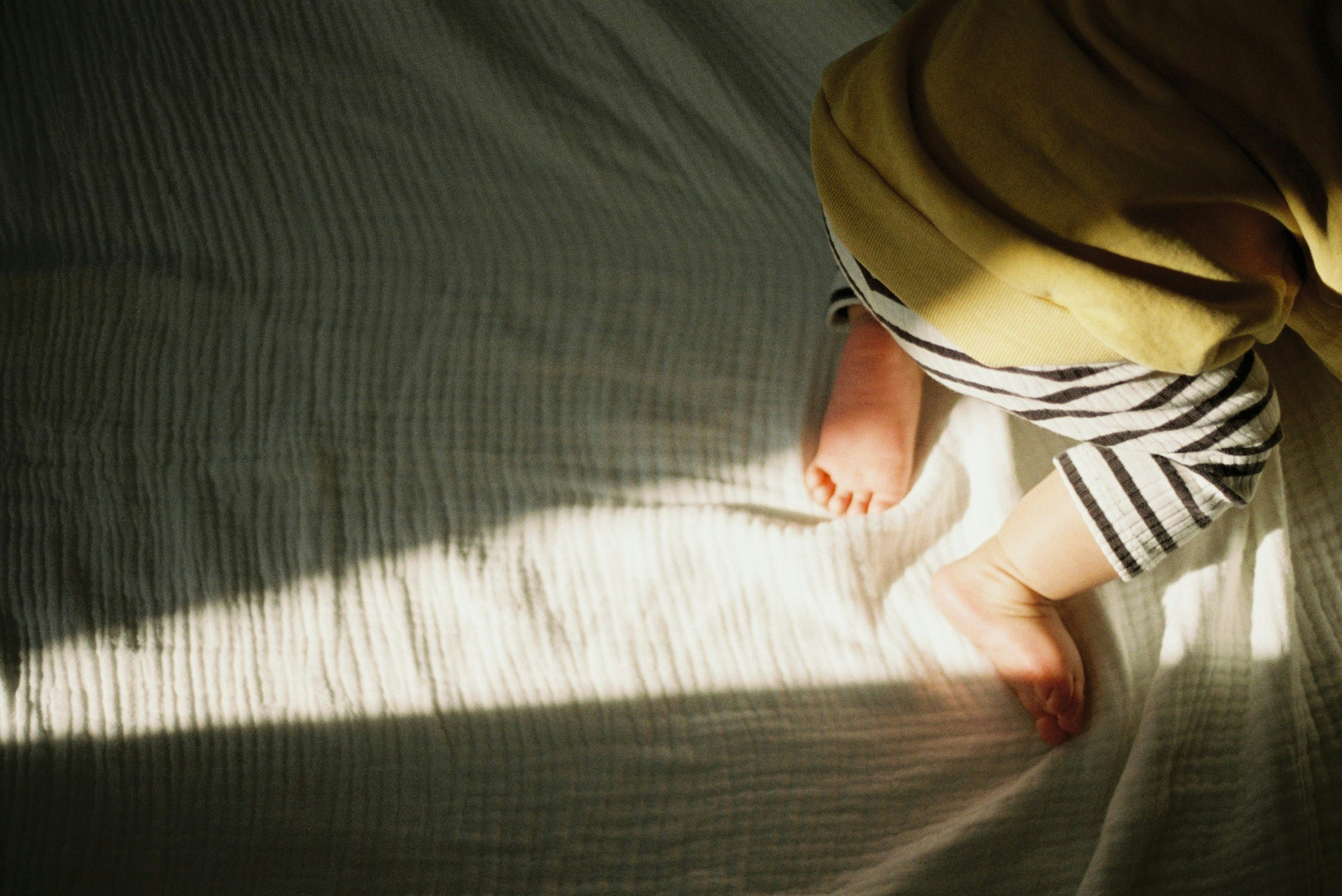 Pies de bebé en pantalones a rayas descansando sobre una cama con luz suave proyectando sombras