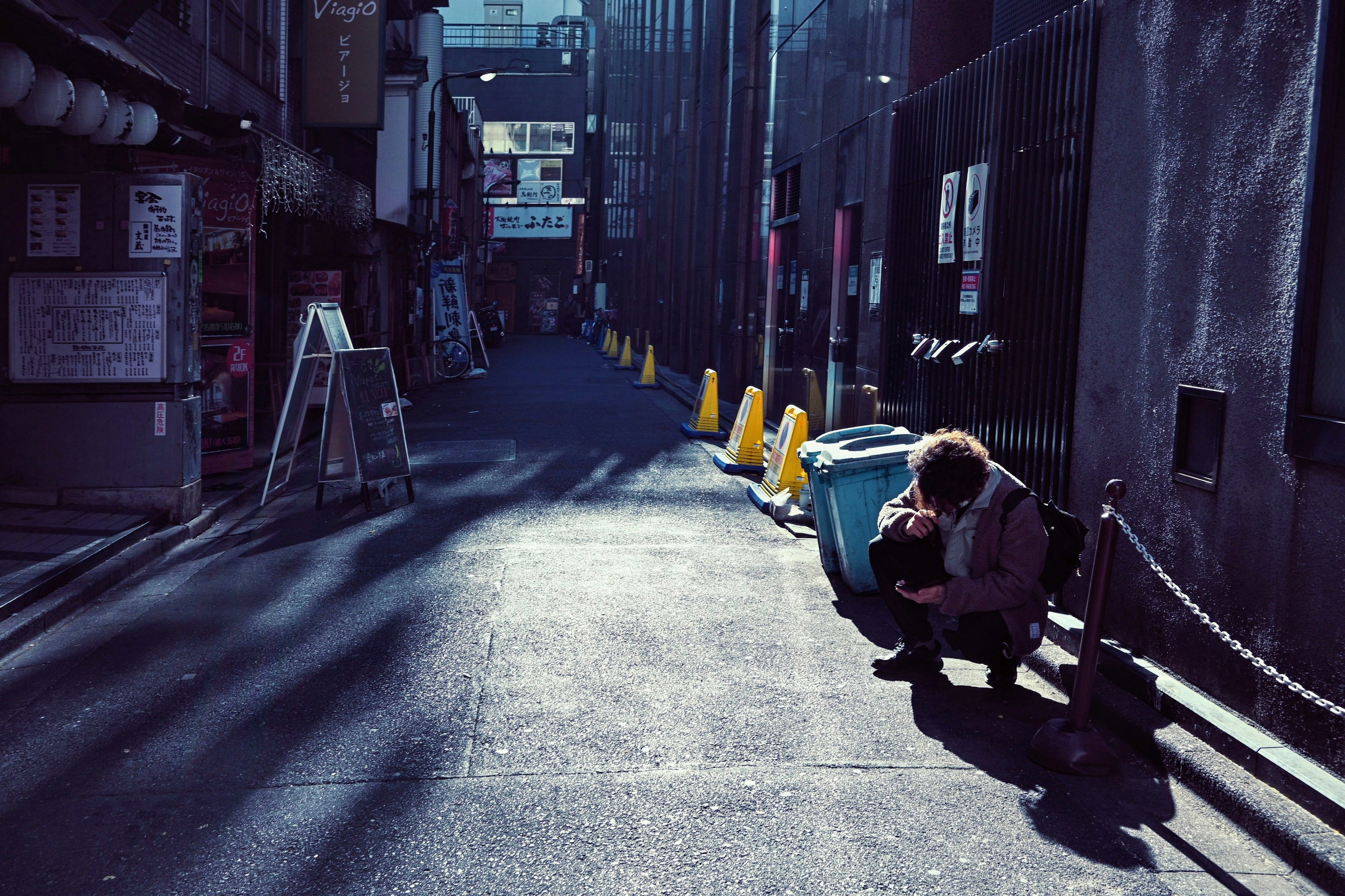 A person crouching in a narrow alley with a blue trash can