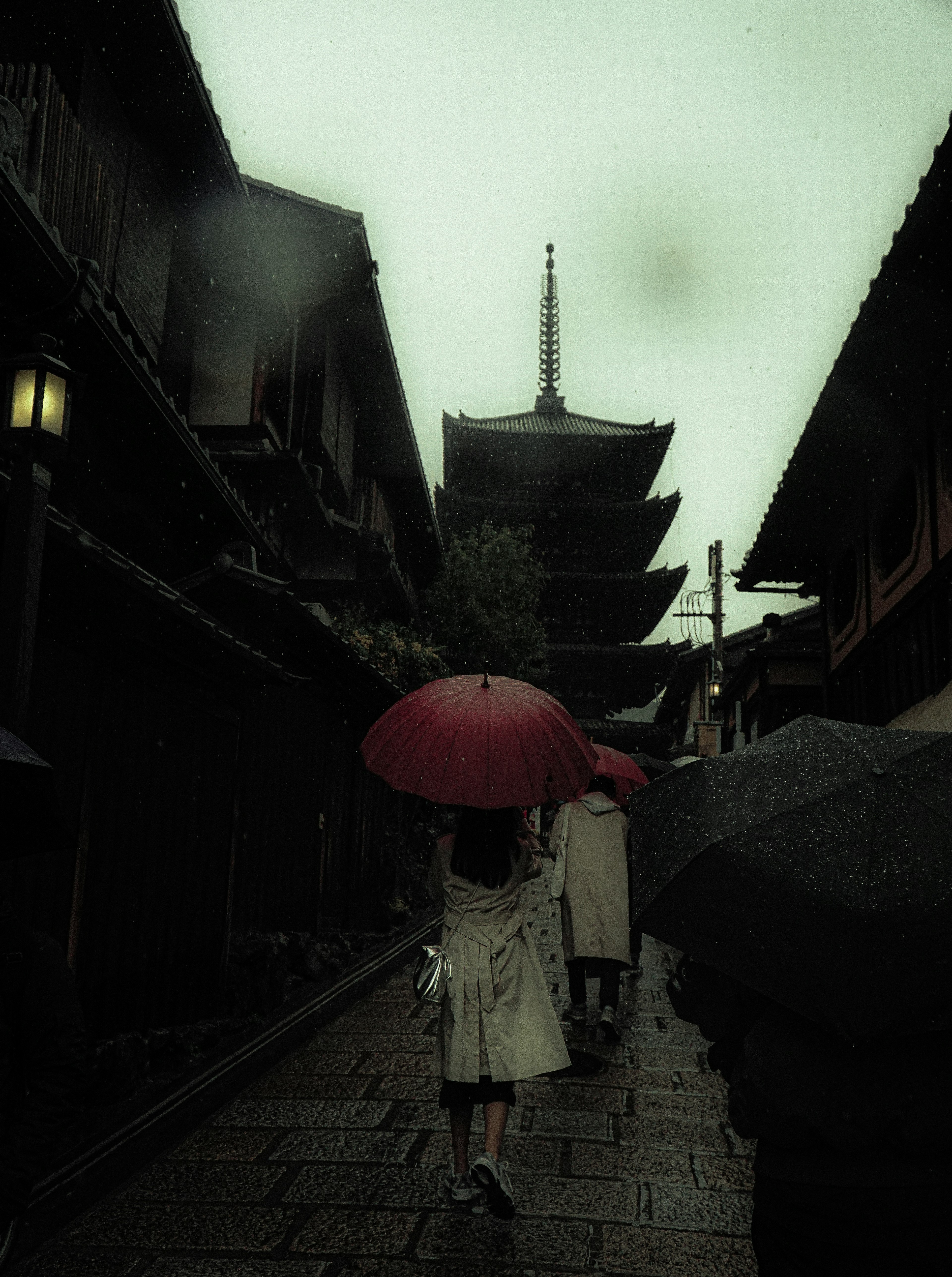 Eine Frau mit einem roten Regenschirm geht durch eine alte Straße mit einer Pagode im Hintergrund an einem regnerischen Tag