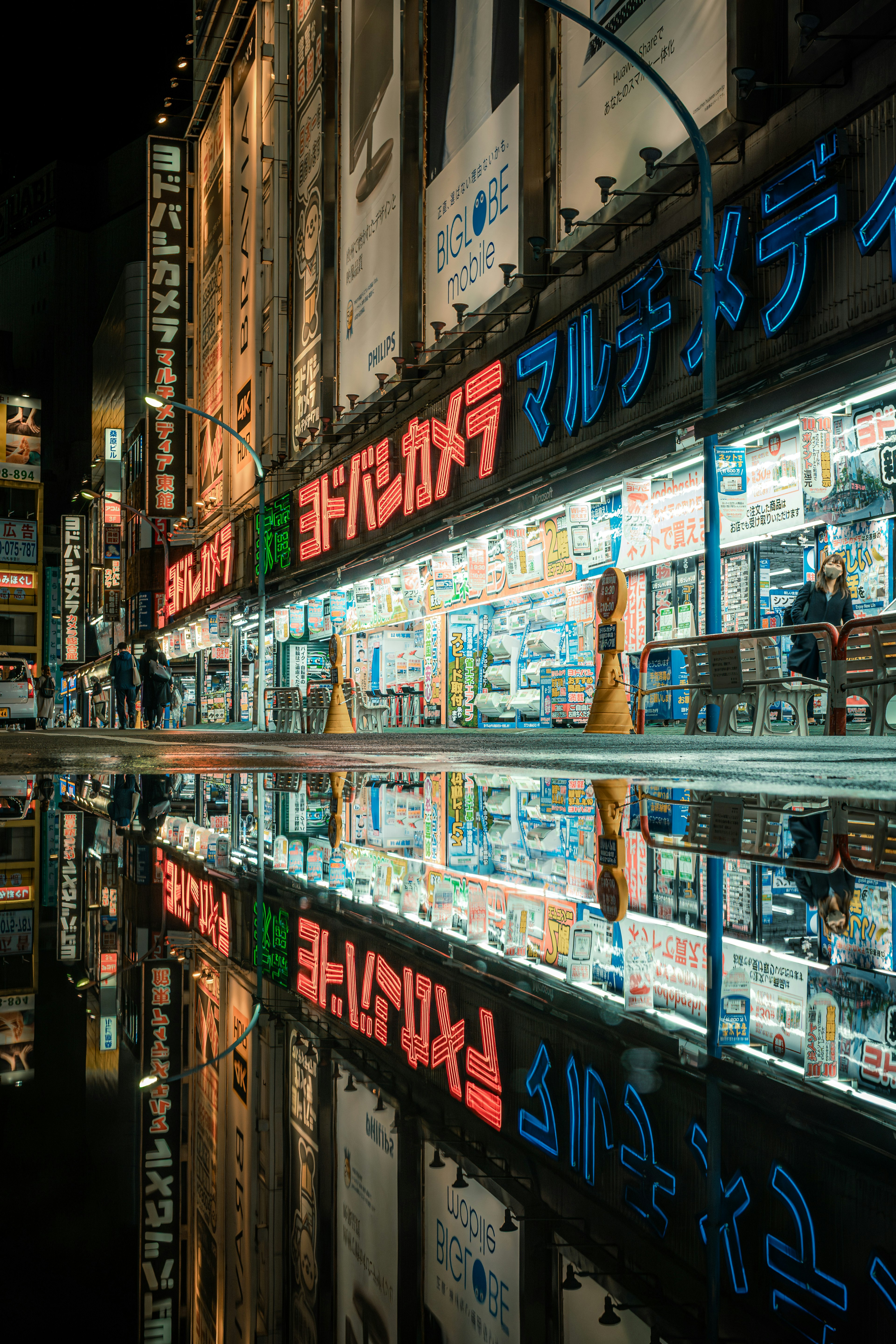 Neon signs and storefronts reflected in a puddle with a person walking