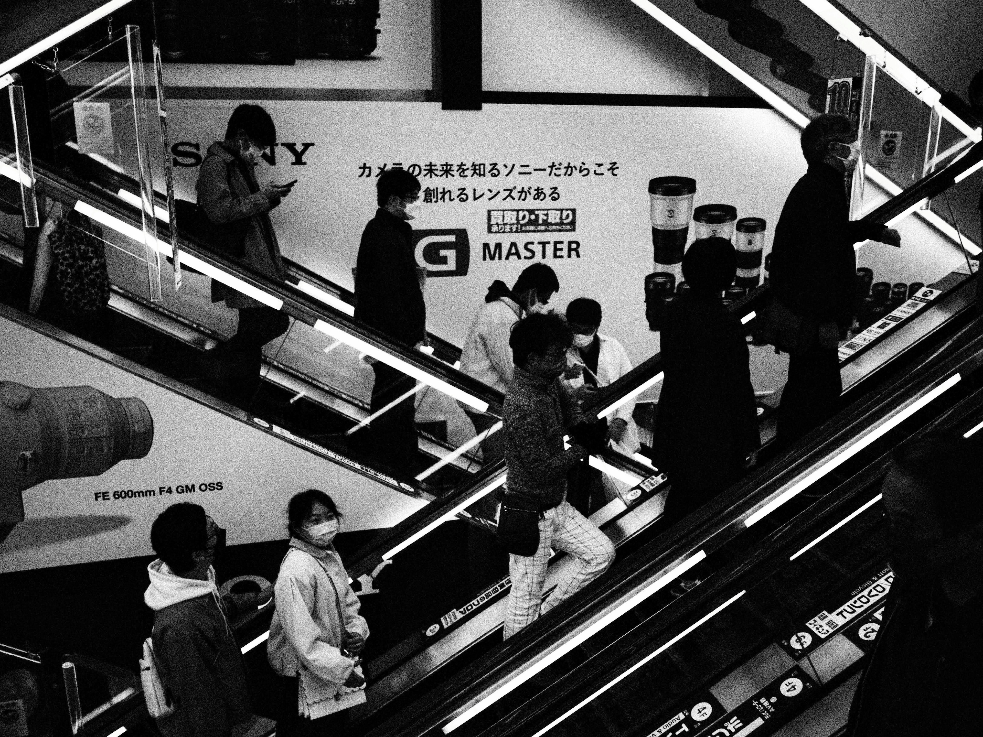 Imagen en blanco y negro de personas utilizando una escalera mecánica con un anuncio de Sony al fondo