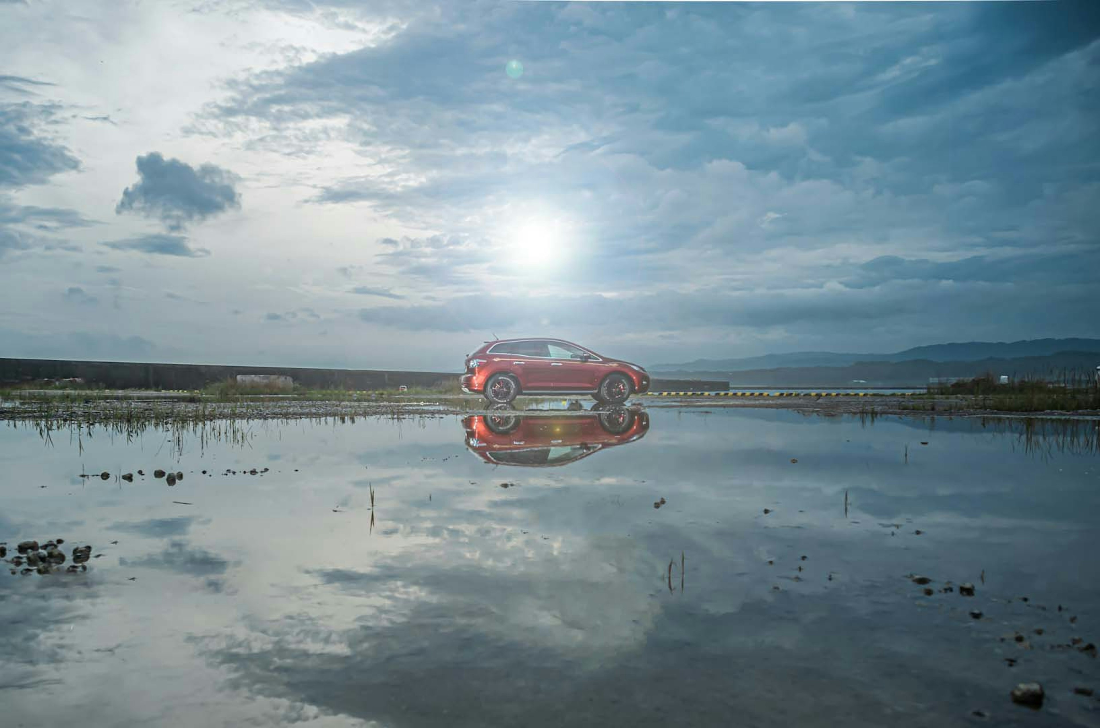 Un'auto rossa riflessa in una superficie d'acqua sotto un cielo blu con nuvole