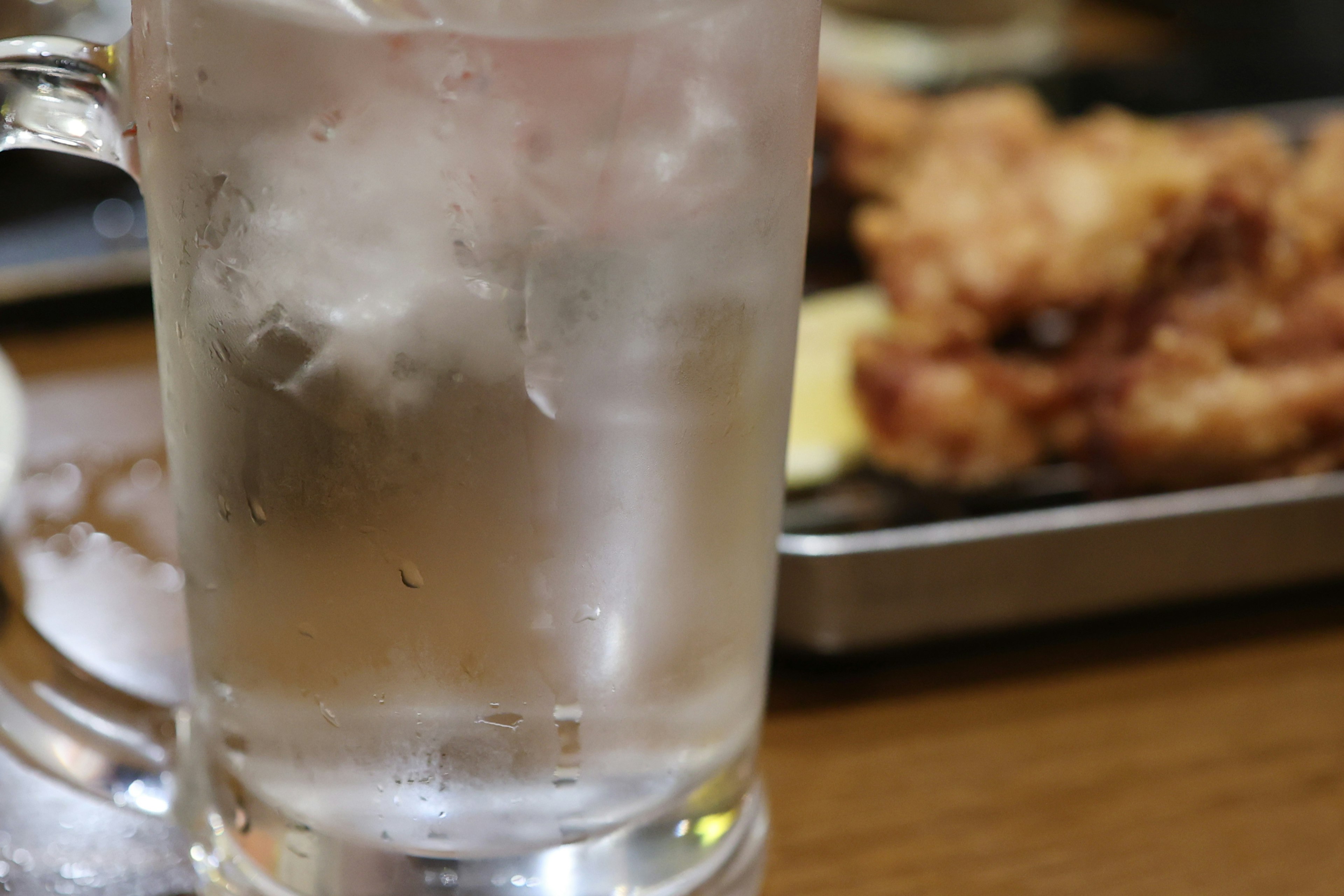 Un vaso transparente lleno de hielo y un plato de comida frita al fondo