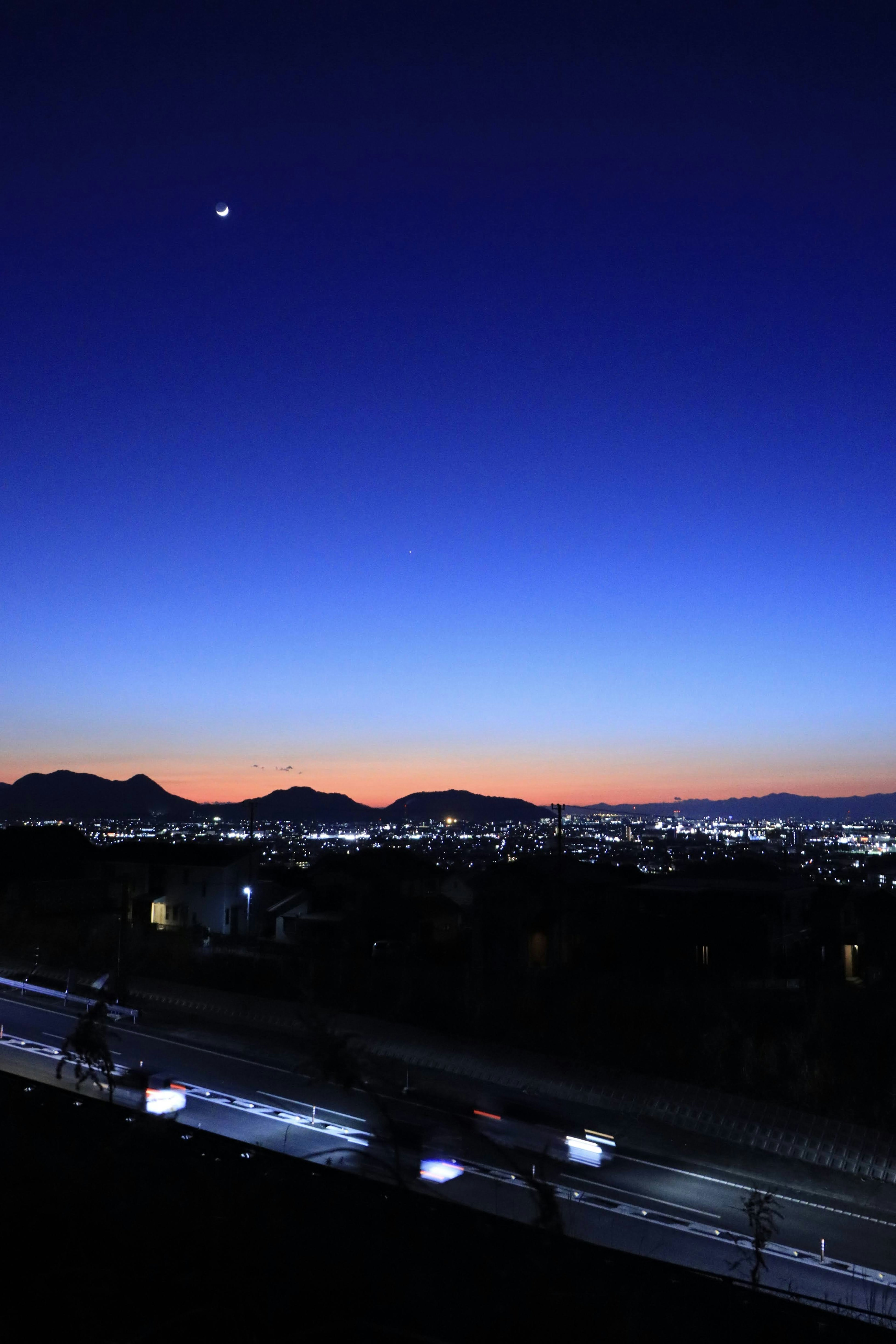 夕焼けの空に浮かぶ月と都市の明かりが映る風景