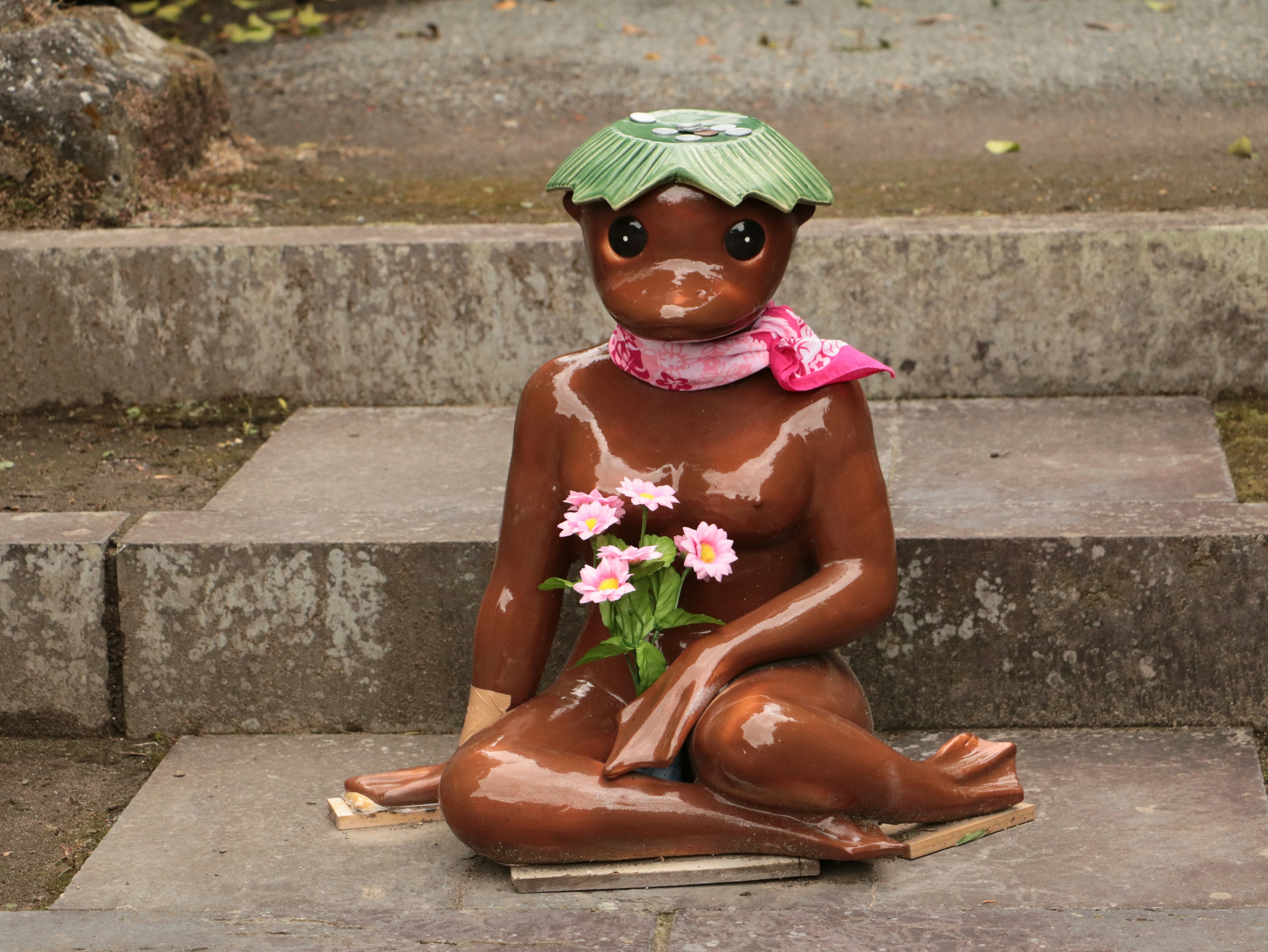 Une statue en céramique portant un chapeau en feuille verte et une écharpe rose tenant des fleurs en position assise