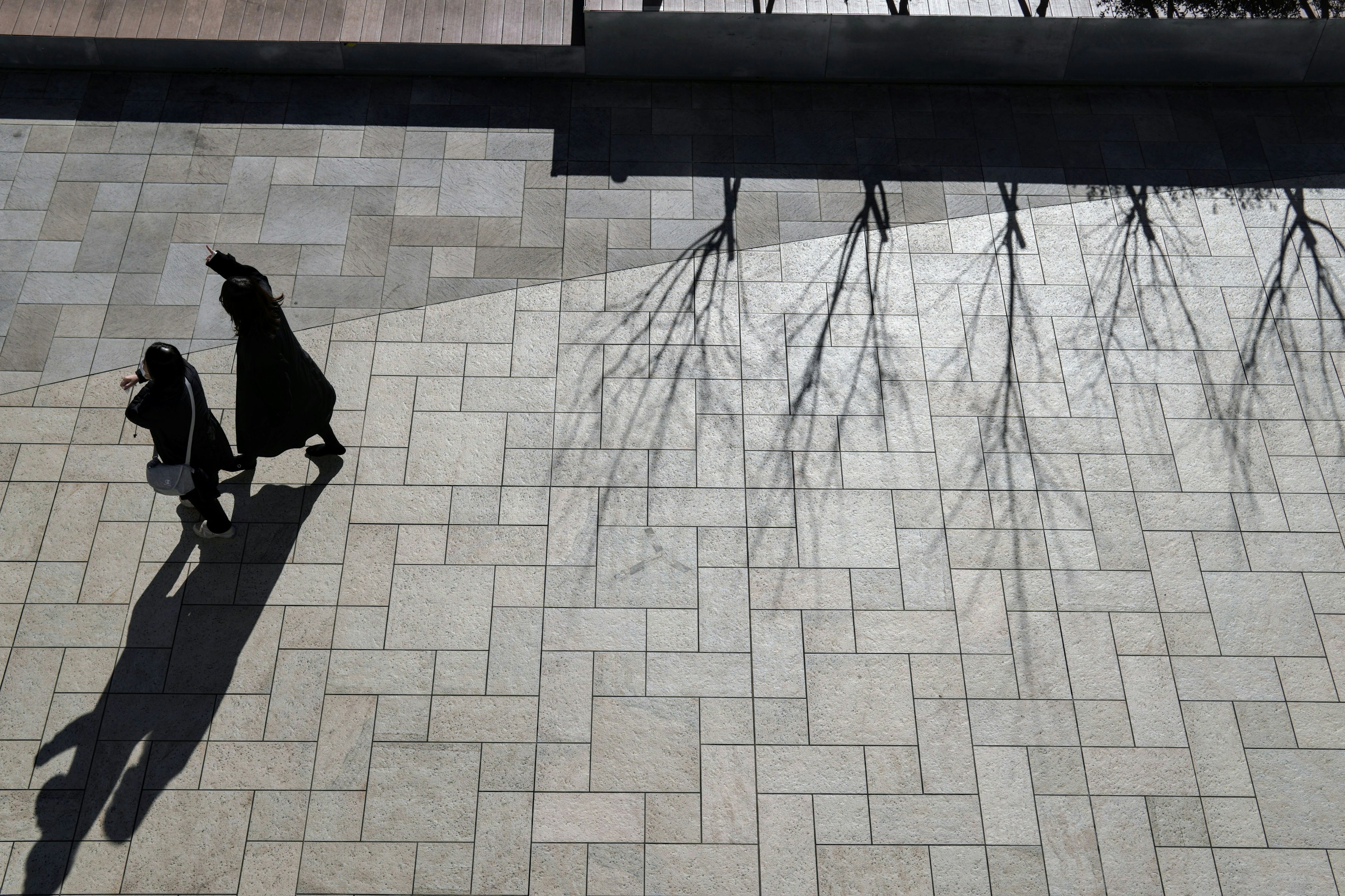 Two figures walking on a stone pavement casting shadows